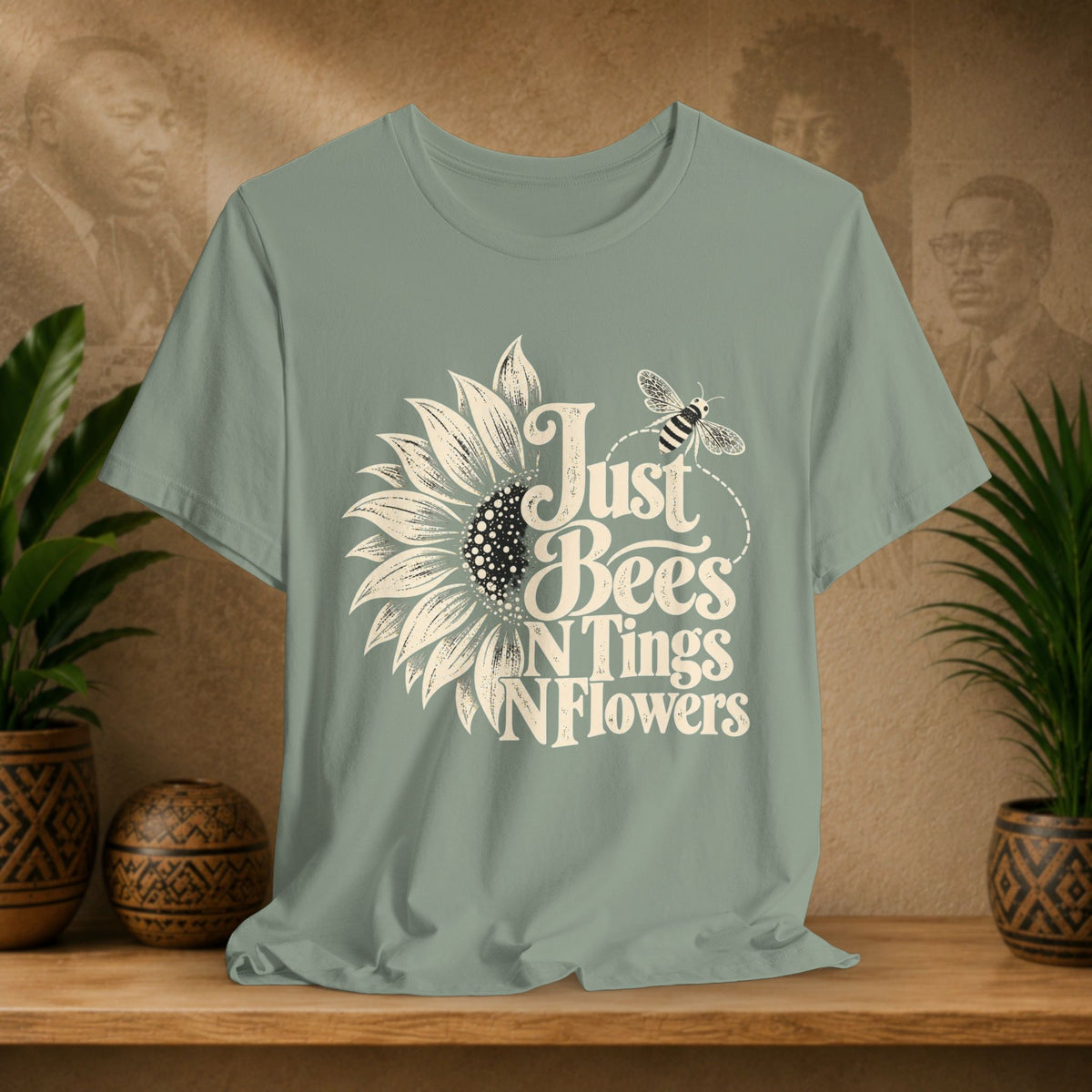 A sage green short-sleeve t-shirt with a 'Just Bees N Tings N Flowers' sunflower and bee graphic, displayed on a wooden shelf with potted plants and a rustic background.