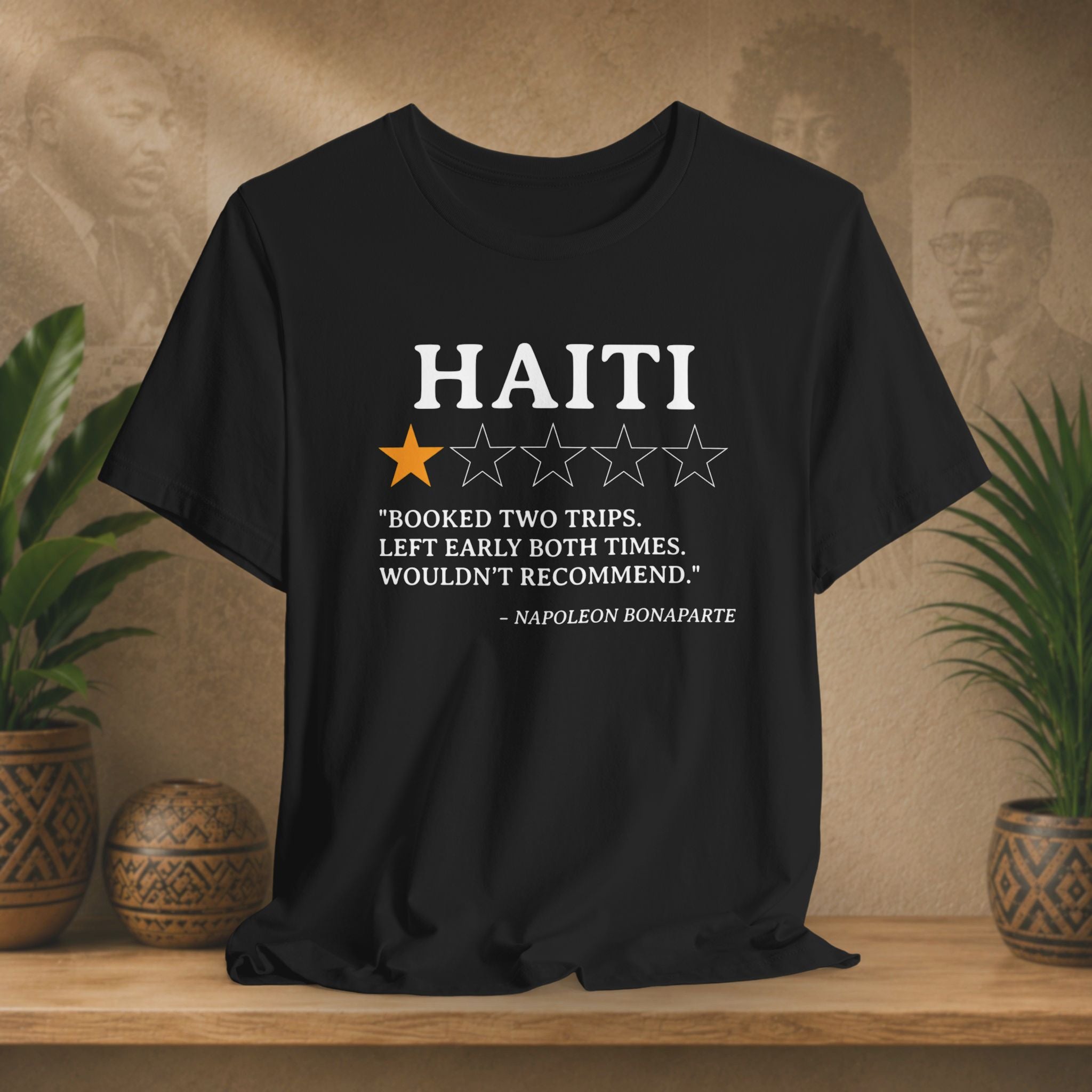 Black t-shirt with Haiti rating and Napoleon Bonaparte quote on a wooden surface with plants and wall art.