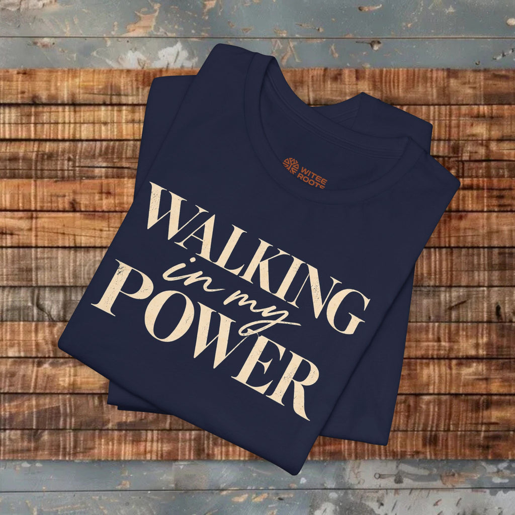A navy blue folded t-shirt with the phrase 'WALKING in my POWER' printed in a cream-colored serif and script font, displayed on a rustic wooden plank background.