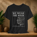 Black t-shirt with 'We Wear the Mask' quote by Paul Laurence Dunbar on a wooden shelf with plants and decorative items.