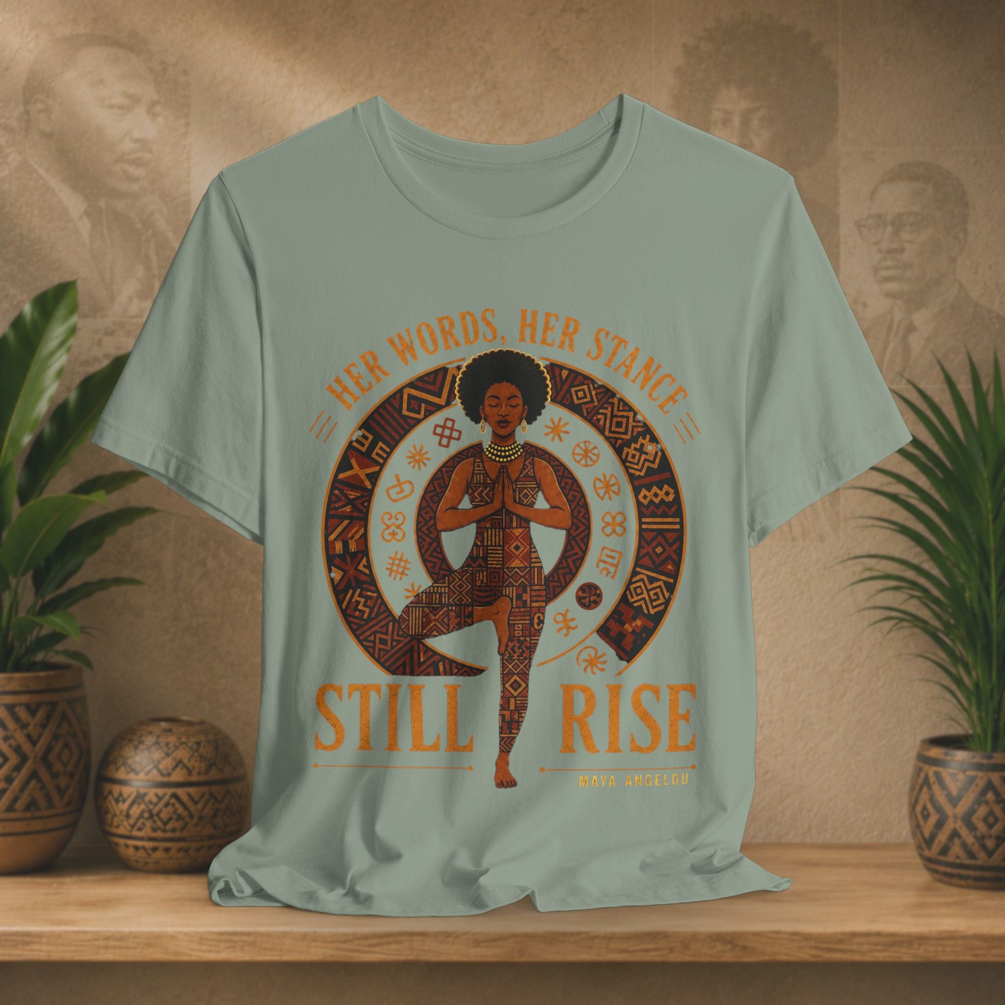 A sage green graphic t-shirt displayed on a wooden shelf with potted plants. The shirt features a circular graphic of a woman in a yoga pose with the text 'HER WORDS.