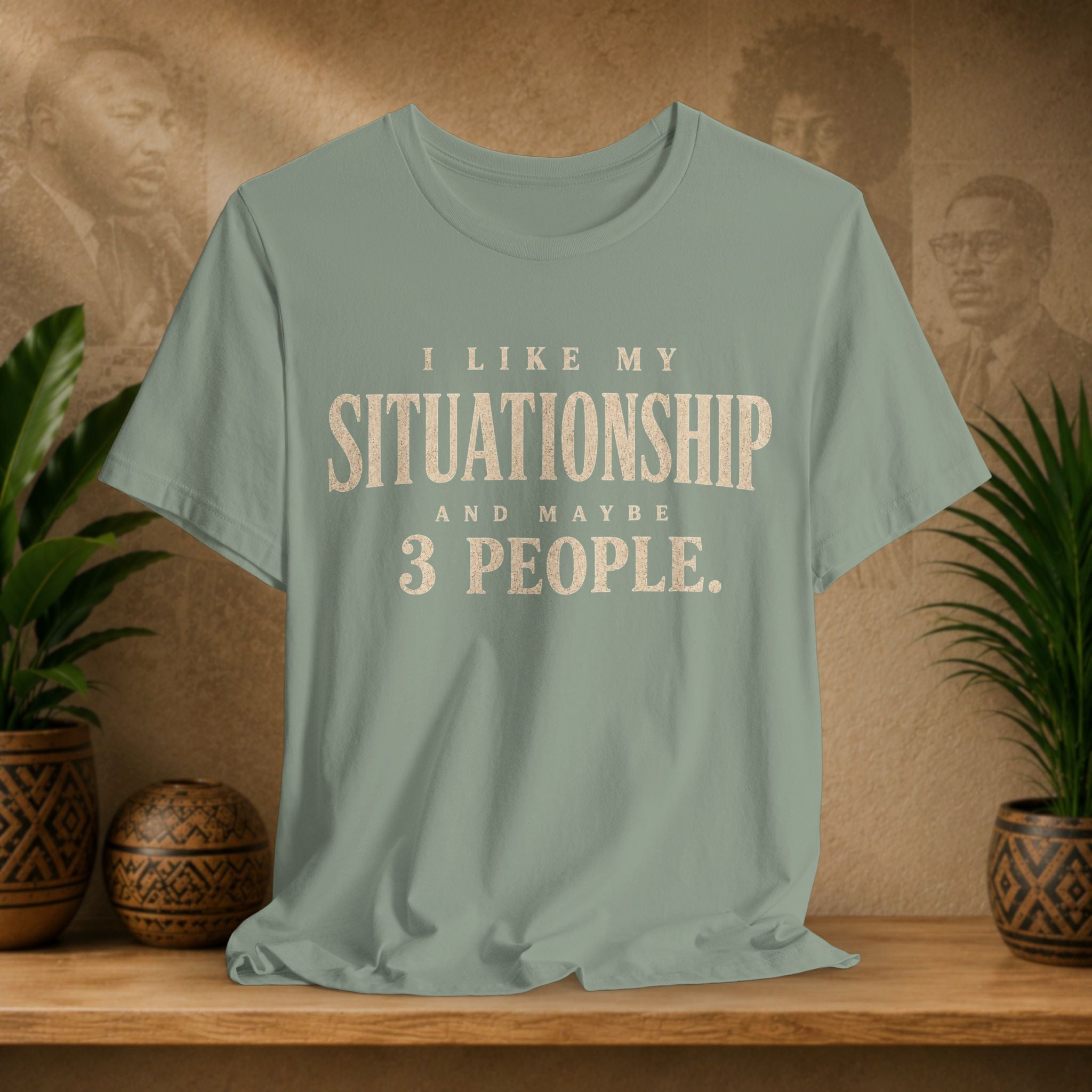 A sage green graphic t-shirt displayed on a wooden shelf, flanked by potted green plants against a textured brown background with vintage portraits. The t-shirt features the text 'I LIKE MY SITUATIONSHIP AND MAYBE 3 PEOPLE.