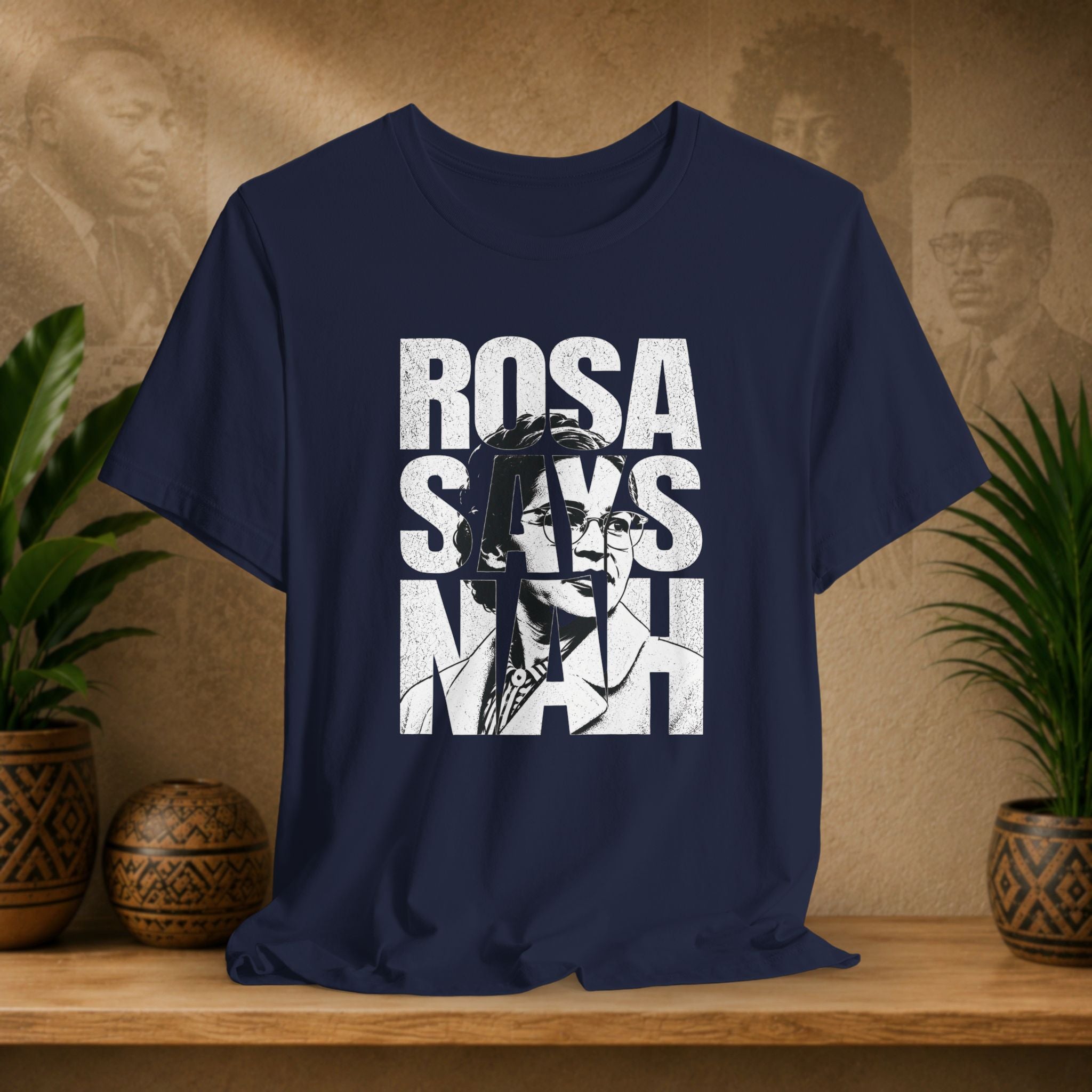Navy blue t-shirt with 'Rosa Parks' graphic and text on a wooden surface with plants and a textured wall background.