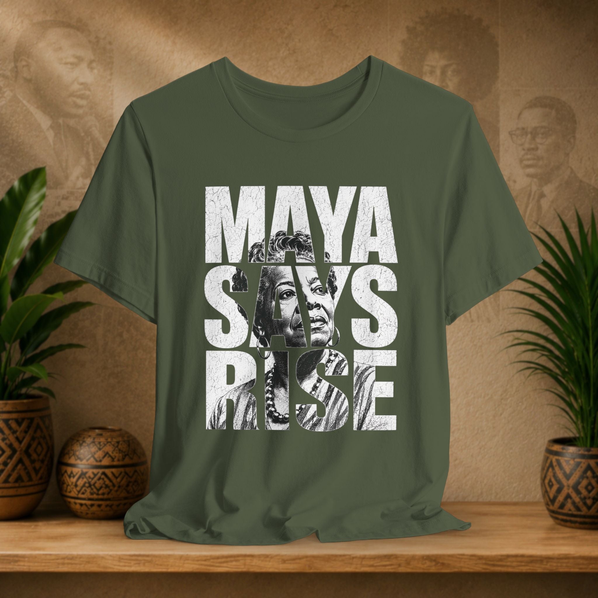 Green t-shirt with 'MAYA SAYS RISE' text and graphic on a wooden surface with plants and a textured wall background.