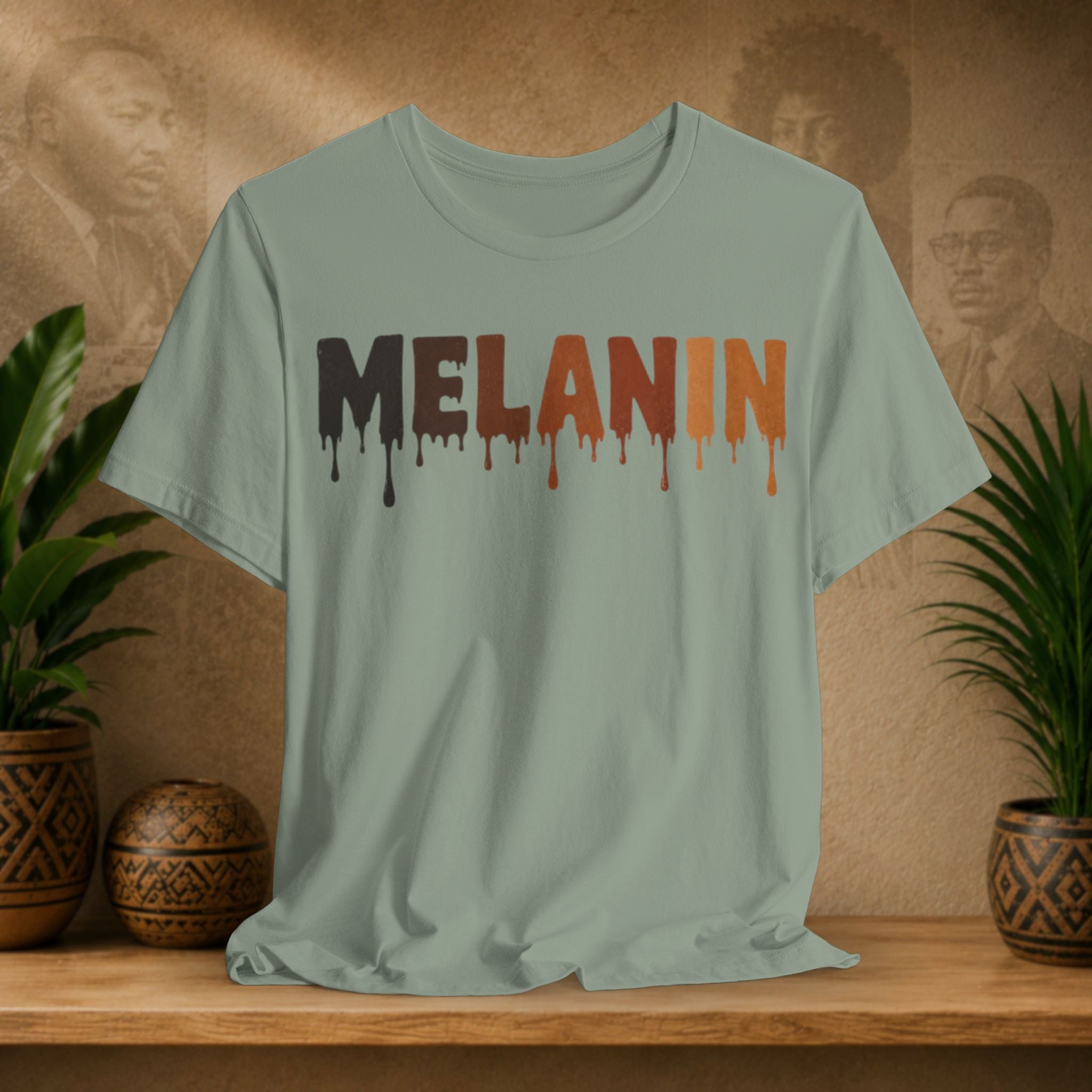 A sage green crewneck t-shirt with the word 'MELANIN' printed in a dripping paint style across the chest. The letters are dark brown/black on the left and orange on the right, with matching dripping paint below.