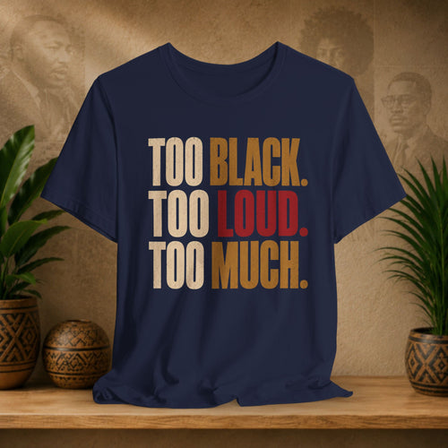 A navy blue graphic t-shirt displayed on a wooden shelf, flanked by potted green plants and decorative ceramic pots against a textured beige wall with sepia portraits. The shirt features the text 'TOO BLACK.