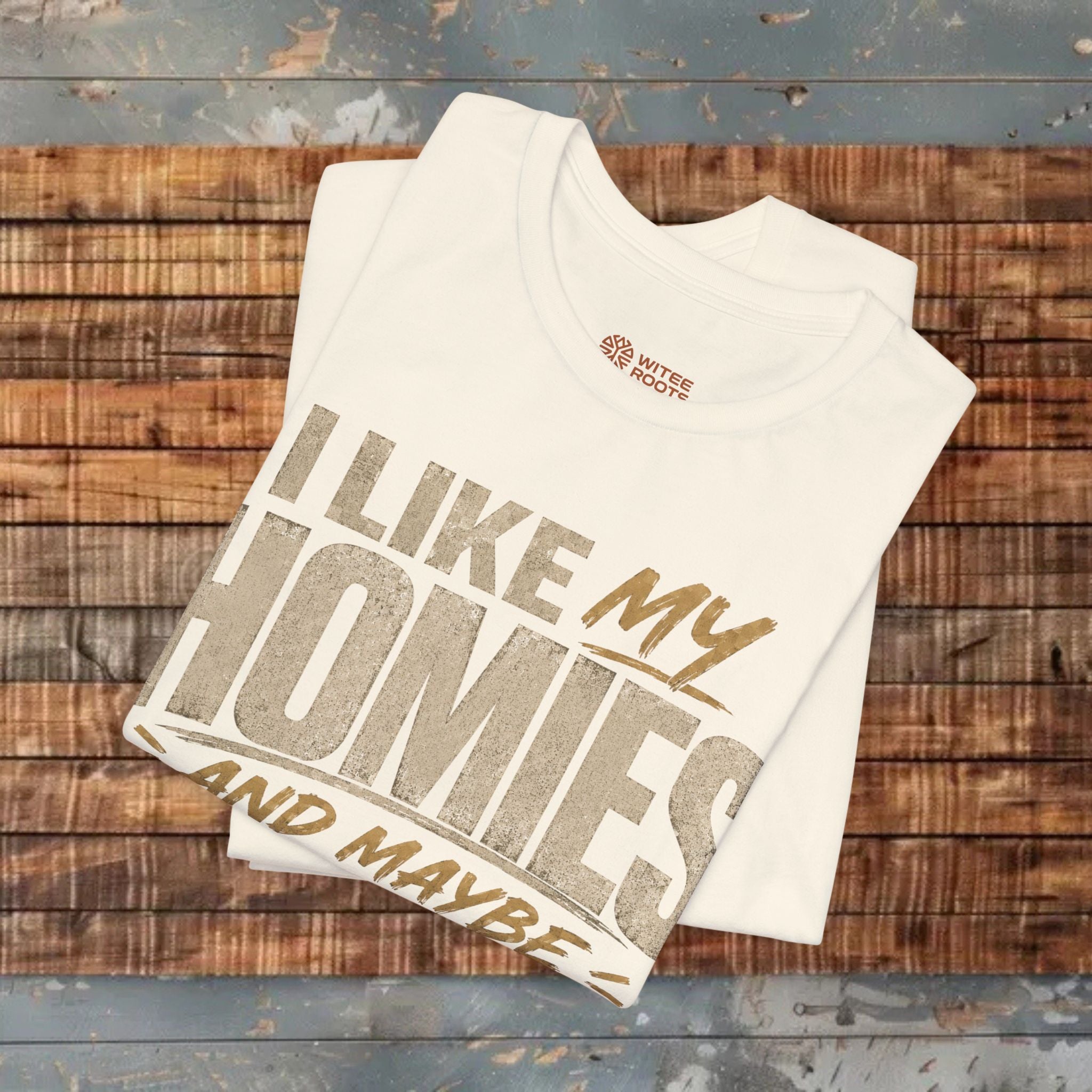 A beige graphic t-shirt folded on a wooden background. The shirt features a humorous slogan printed in white and tan lettering: 'I LIKE MY HOMIES and maybe 3 PEOPLE.
