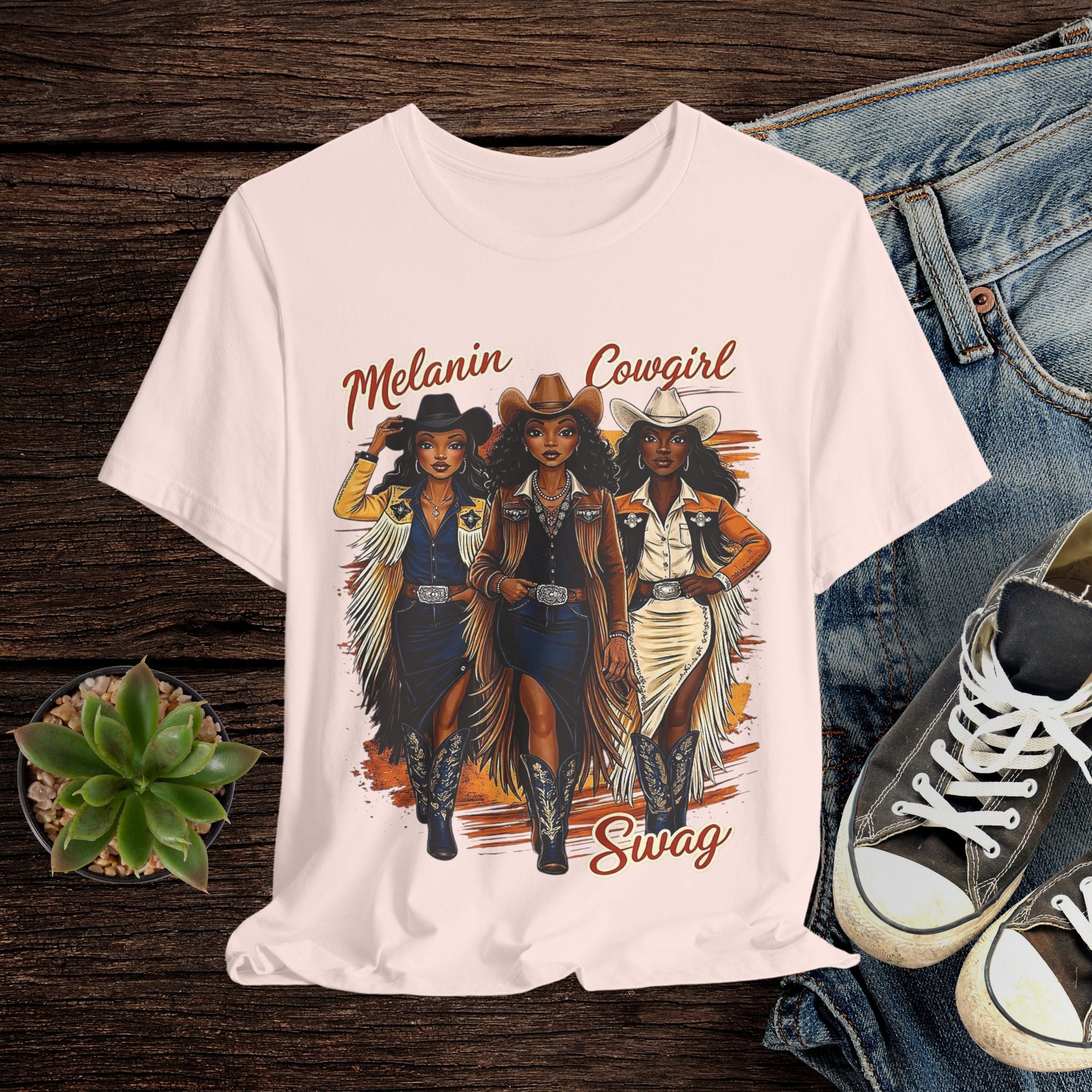 Pink t-shirt with 'Melanin Cowgirl Swag' design featuring three cowgirls on a wooden surface with jeans and sneakers.