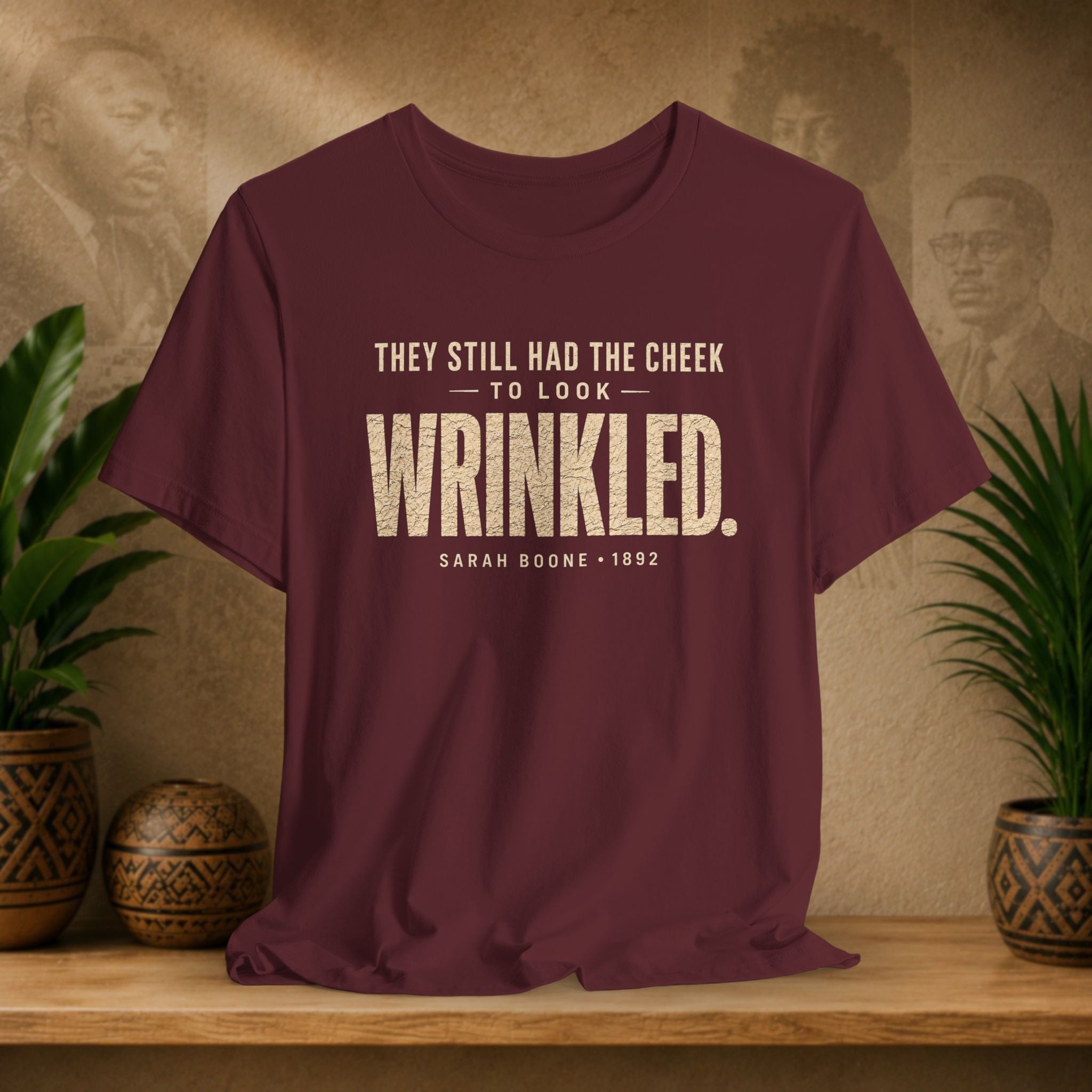Maroon t-shirt with white text on a wooden surface with plants and a textured wall background