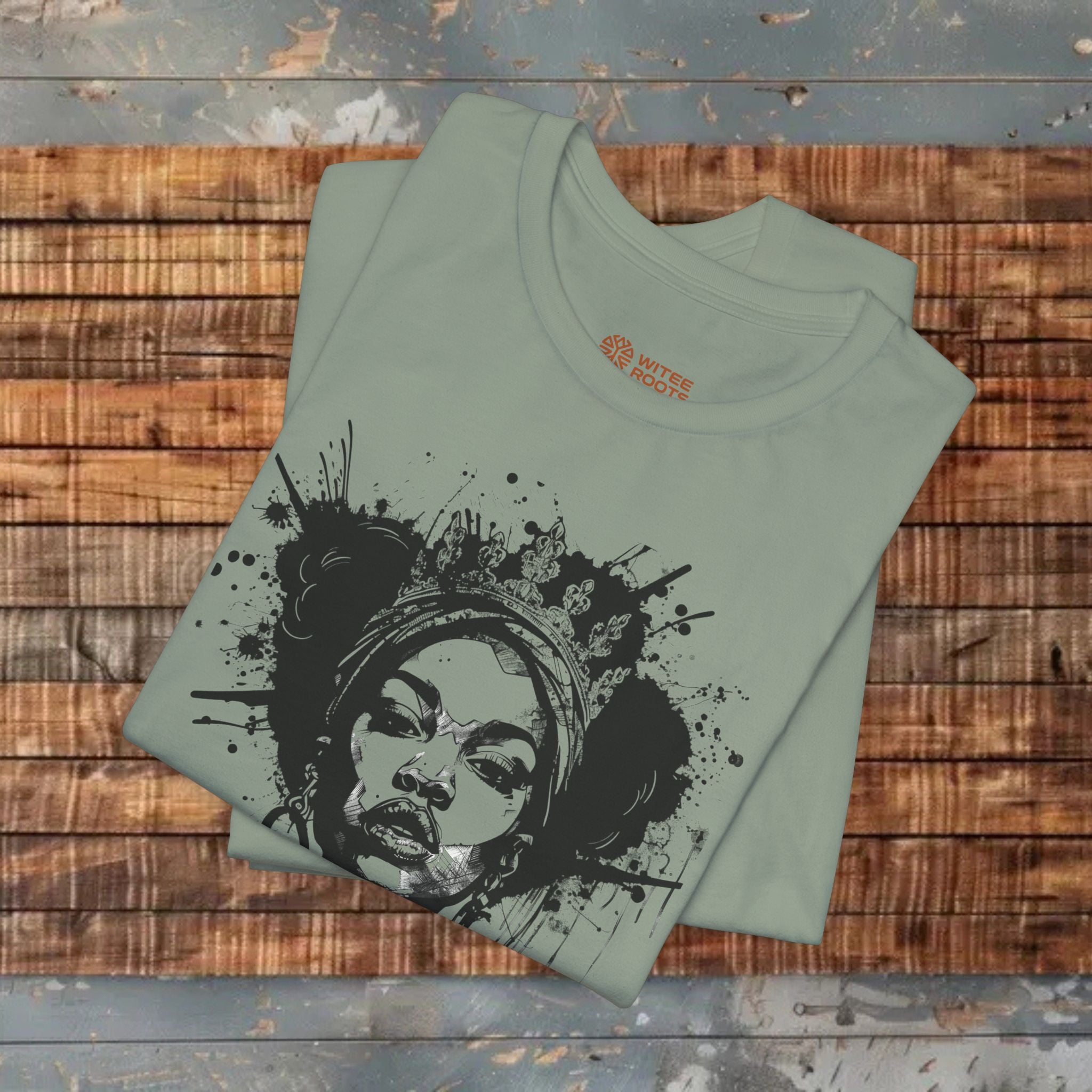 A folded sage green t-shirt with a black and white graphic print of a woman's face surrounded by paint splatters, displayed on a rustic wooden plank background.