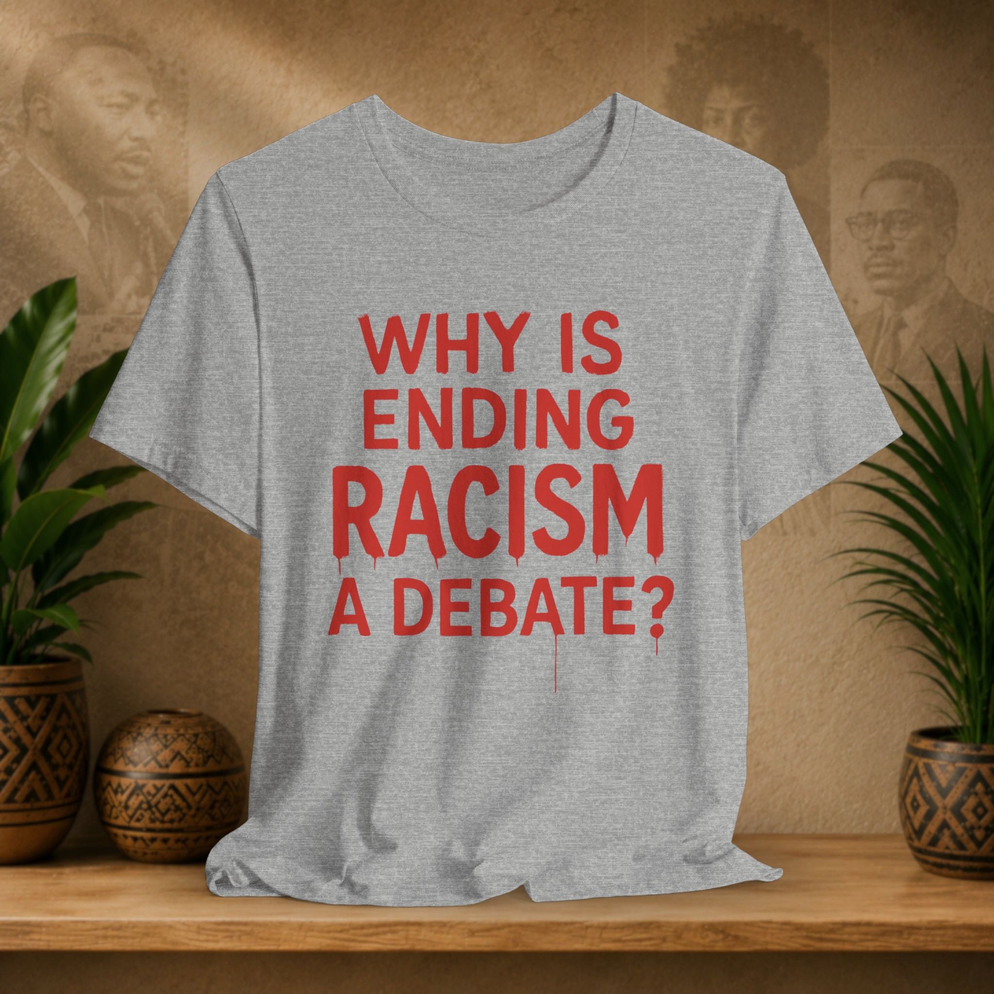 Gray t-shirt with red text 'Why is ending racism a debate?' on a wooden surface with plants and a blurred background.
