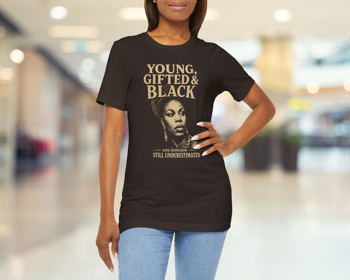A woman modeling a brown graphic t-shirt in a blurred indoor mall setting. The shirt features a gold and beige print with the text 'YOUNG, GIFTED & BLACK' and a portrait of Oprah Winfrey.
