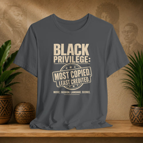 Gray t-shirt with 'Black Privilege' text on a wooden shelf with plants and decorative items.