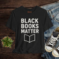 Black t-shirt with 'BLACK BOOKS MATTER' text and book icon on a wooden surface with jeans and sneakers.