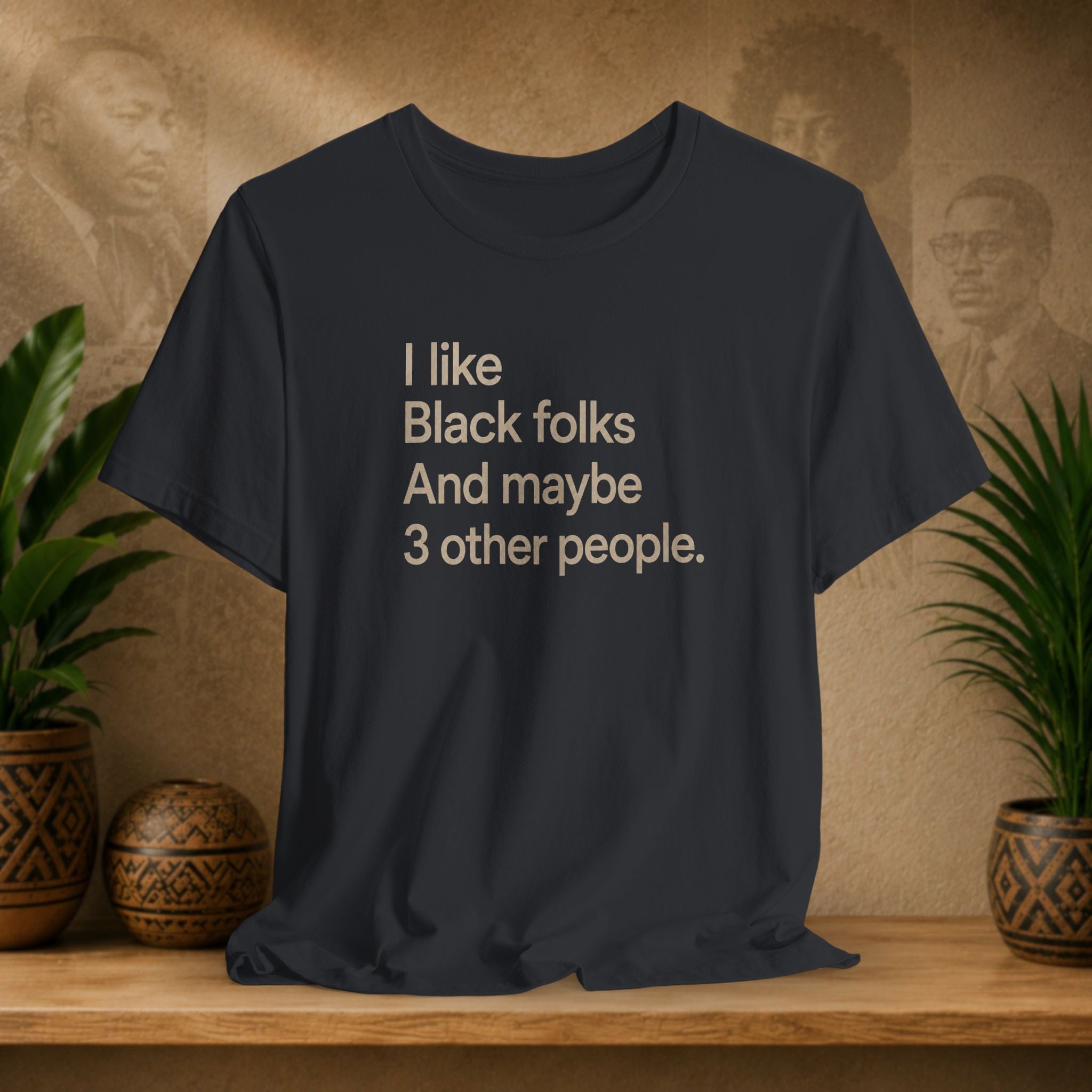 A dark charcoal gray graphic t-shirt displayed on a wooden shelf, flanked by potted green plants and decorative ceramic vases against a textured beige wall with sepia portraits.