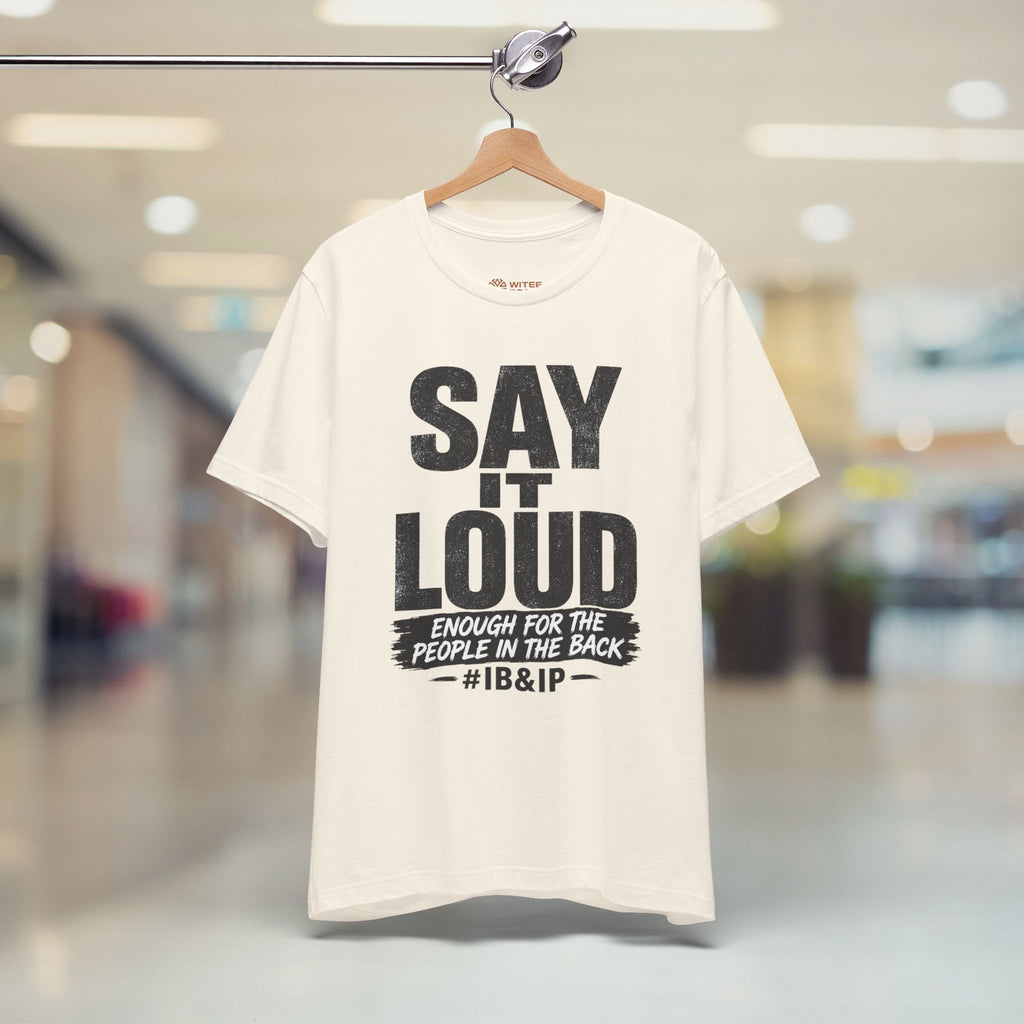 Beige t-shirt with black text on a hanger in a blurred indoor setting