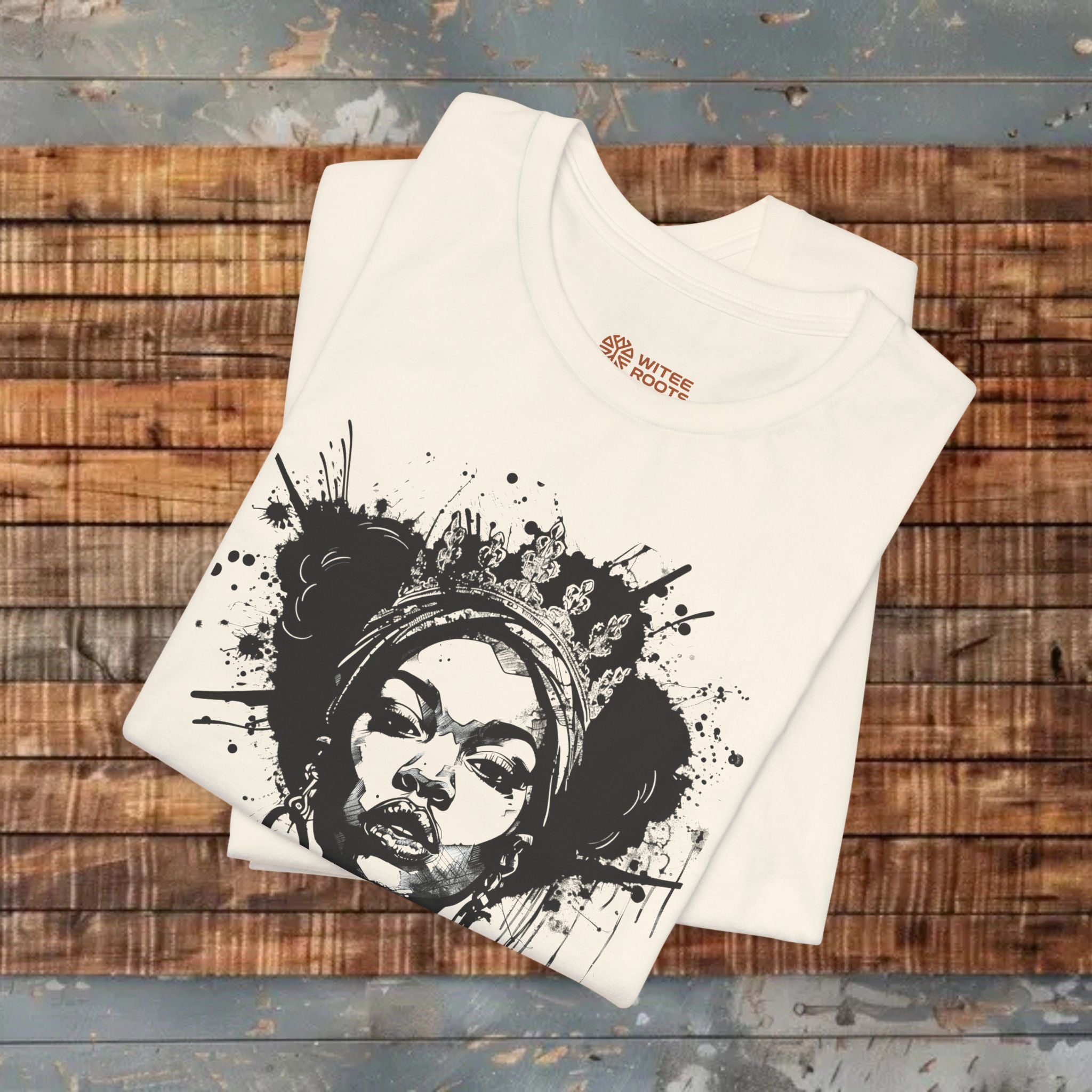 A folded beige t-shirt with a black and white graphic print of a woman's face surrounded by paint splatters, displayed on a rustic wooden plank surface.