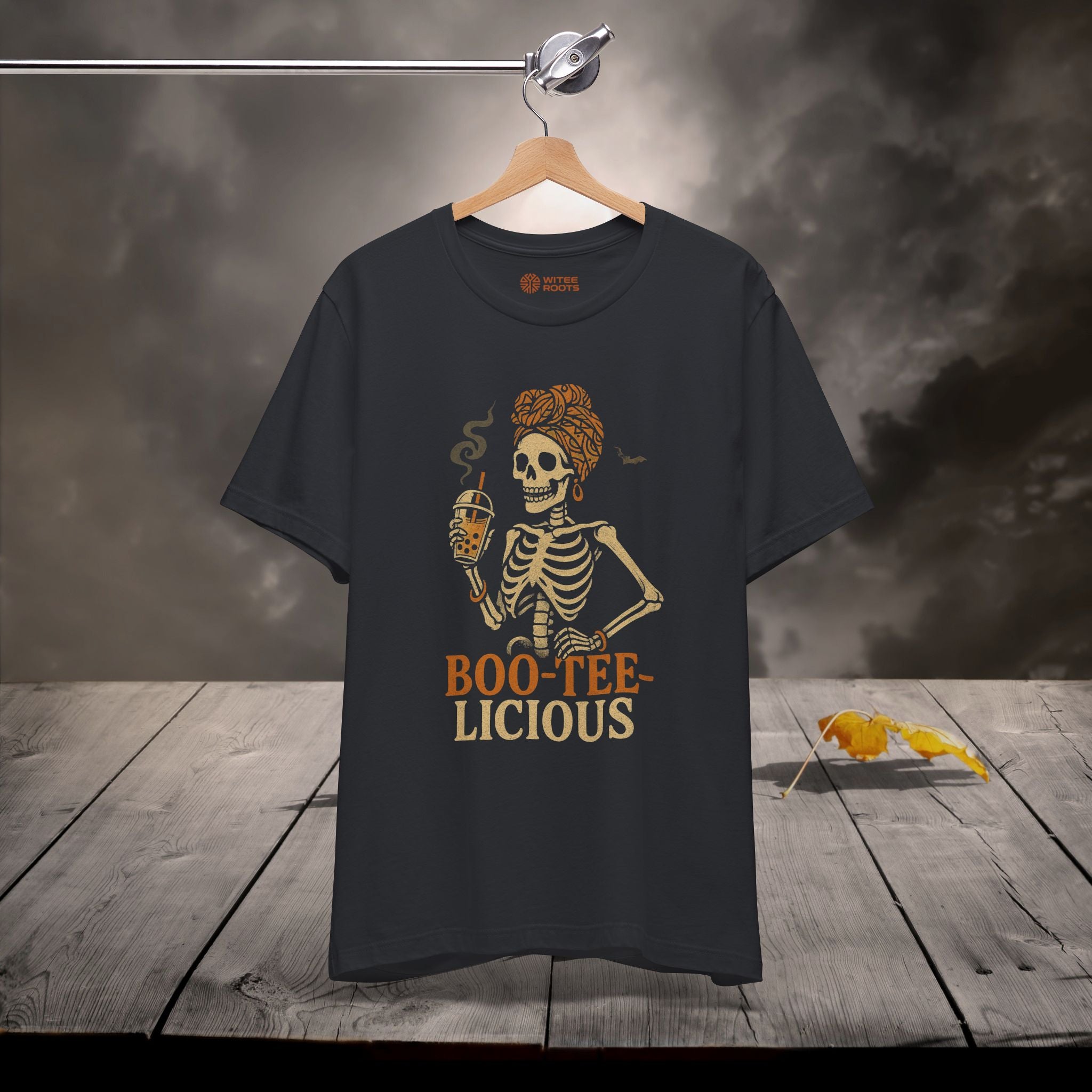 A black graphic t-shirt hanging on a wooden hanger against a moody, smoky background with a rustic wooden table. The t-shirt features a graphic of a skeleton with orange hair holding a drink, with the text 'BOO-TEE-LICIOUS' printed below in orange and beige.