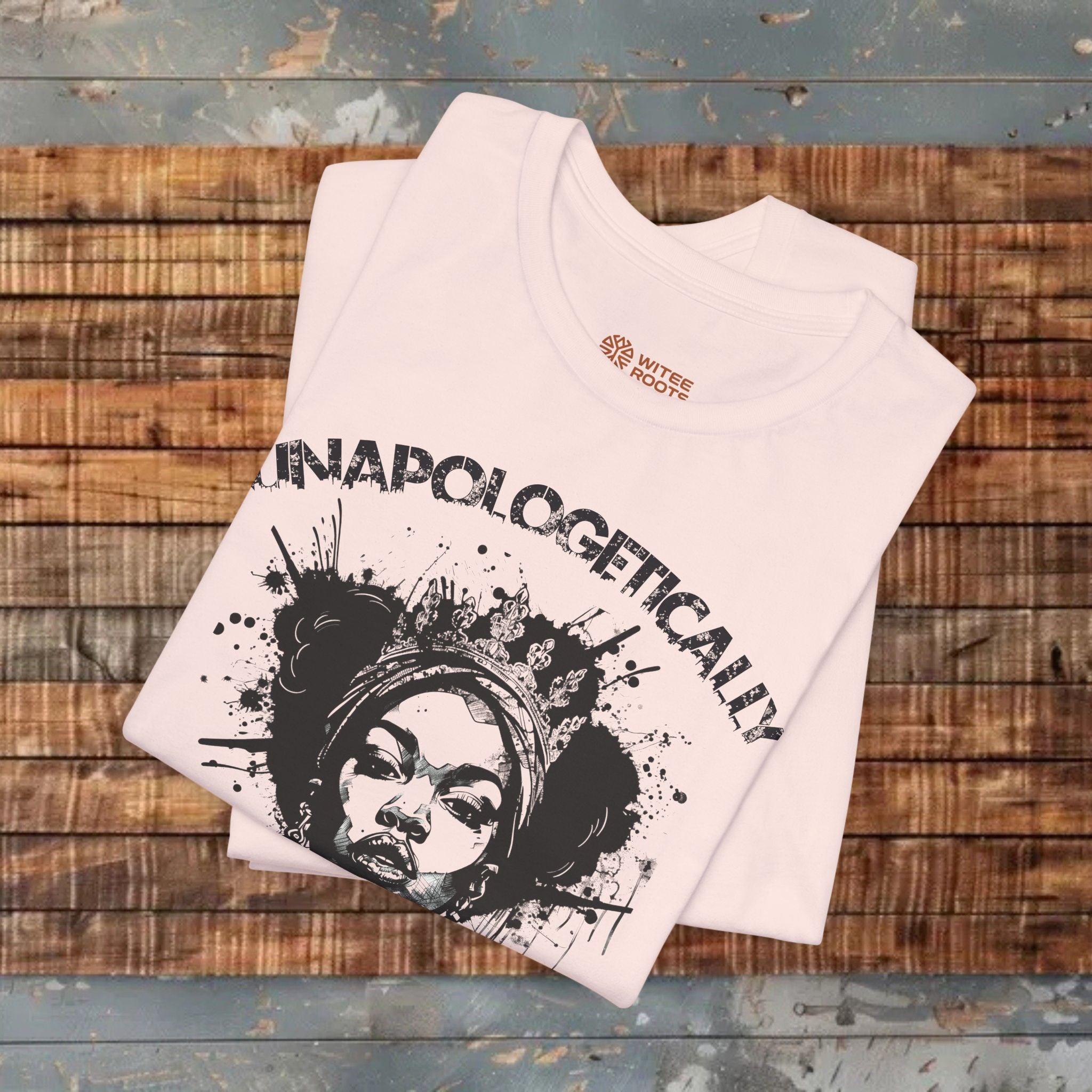 A folded light pink t-shirt with a black graphic print of a woman's face and the word 'UNAPOLOGETICALLY' in arched letters, displayed on a rustic wooden plank background.