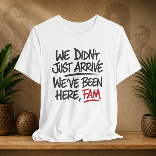 A white short-sleeve graphic T-shirt displayed on a wooden surface, with a warm brown textured background and potted plants on both sides. The shirt features the text 'WE DIDN'T JUST ARRIVE WE'VE BEEN HERE, FAM' in black and red lettering.