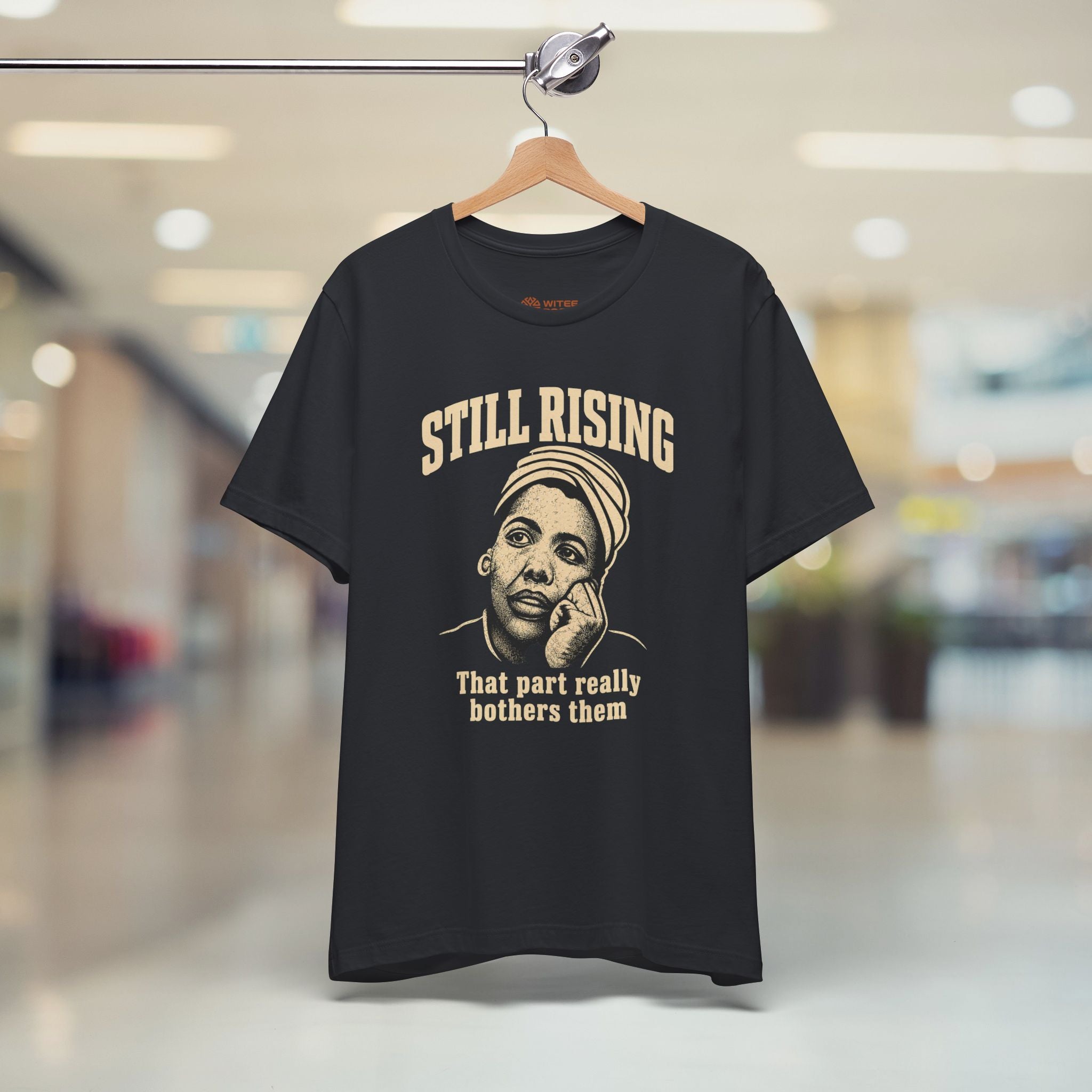 Black t-shirt with 'Still Rising' graphic and text on a hanger in a store setting