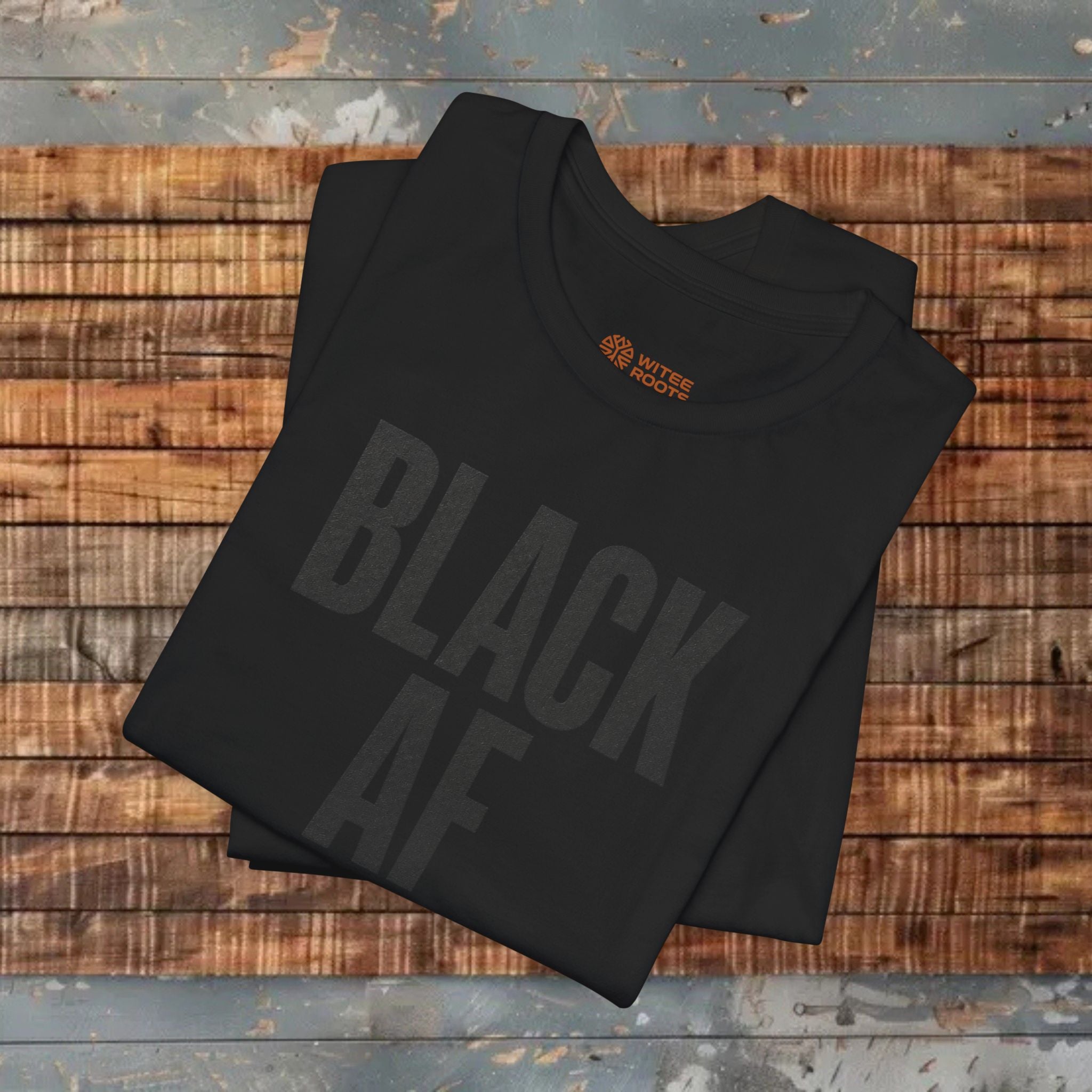 A folded black t-shirt with 'BLACK AF' printed in a faded dark gray font, displayed on a rustic wooden plank surface. The inner collar features a small orange logo for 'WTF WITCHGATE'.