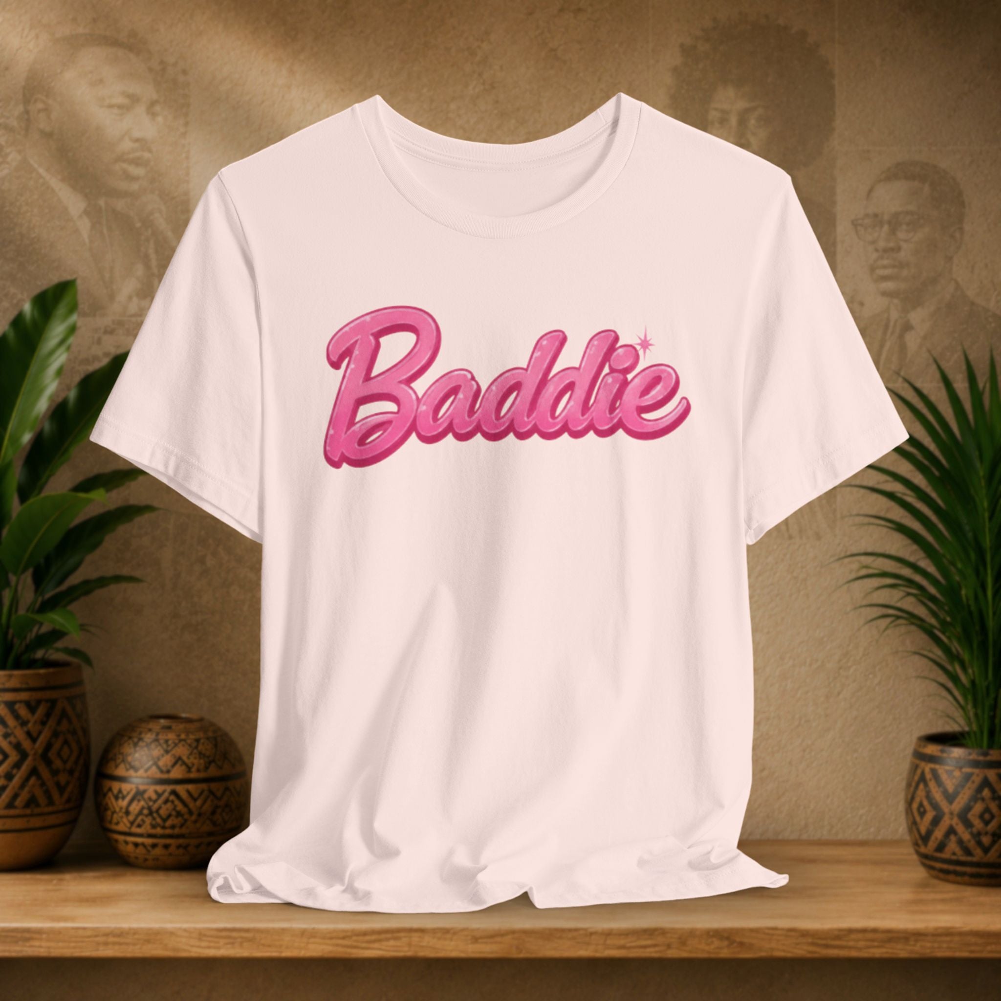 A light pink short-sleeve T-shirt with the word 'Baddie' printed across the chest in a pink retro 3D-style font, displayed on a wooden surface with potted plants and a textured wall background.