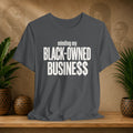 A dark gray t-shirt with white text reading 'minding my BLACK-OWNED BUSINESS$$' displayed on a wooden shelf with potted plants and a textured background.