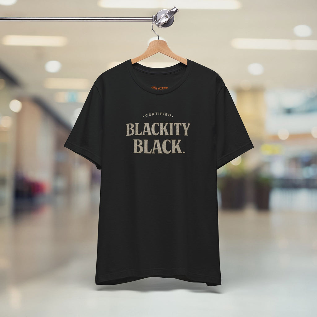 Black t-shirt with 'CERTIFIED BLACKITY BLACK' text on a hanger in a blurred indoor setting