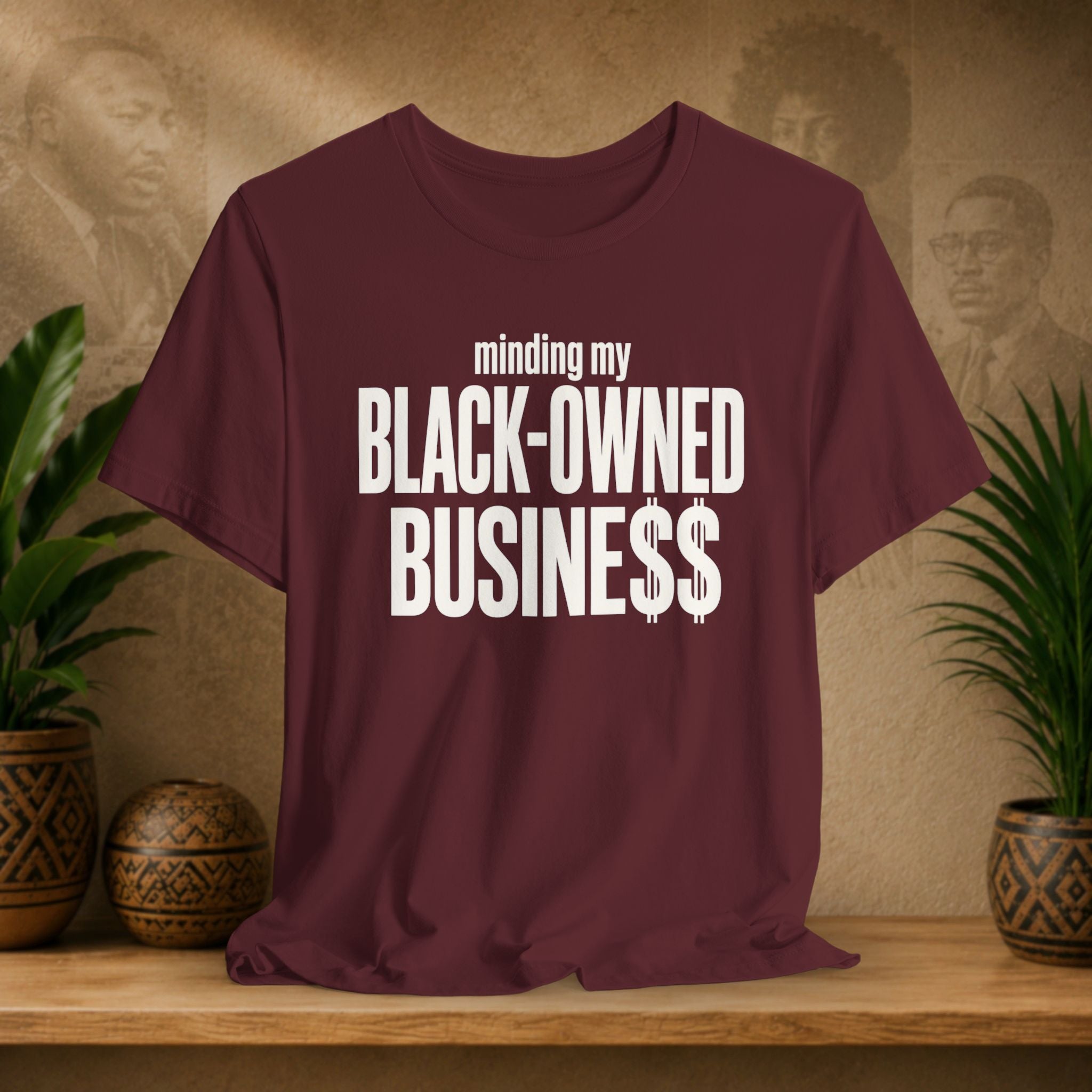 A maroon short-sleeve t-shirt with white text reading 'minding my BLACK-OWNED BUSINESS$$' displayed on a wooden shelf with potted plants and a textured background.