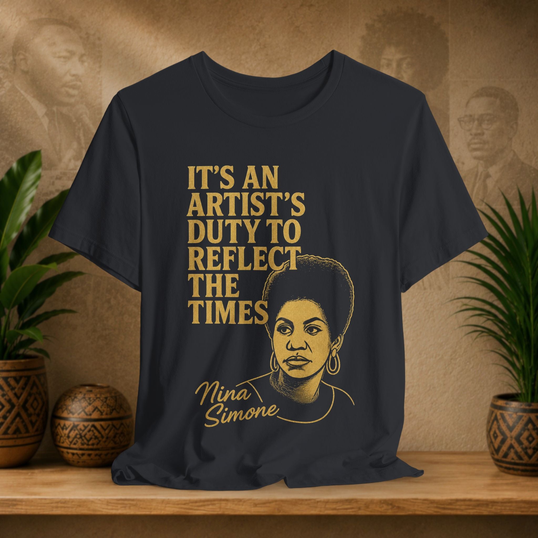 Black t-shirt with gold text and Nina Simone illustration on a wooden surface with plants and wall art in the background.
