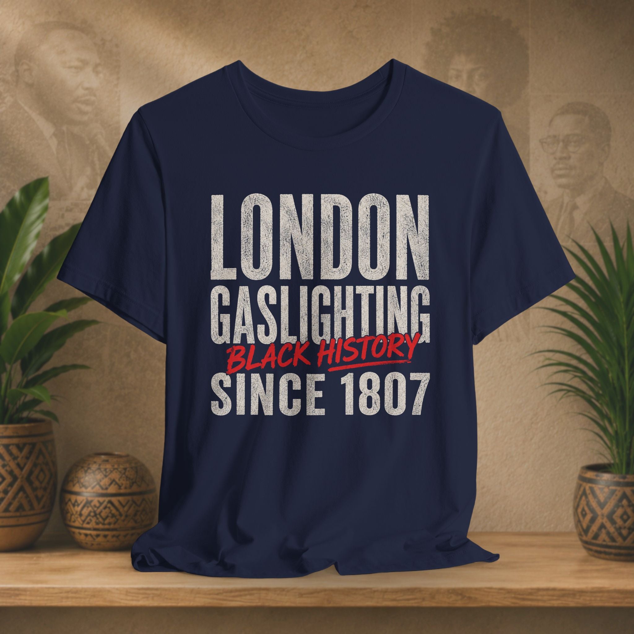 Navy blue t-shirt with text 'LONDON GASLIGHTING BLACK HISTORY SINCE 1807' on a wooden surface with plants in the background.