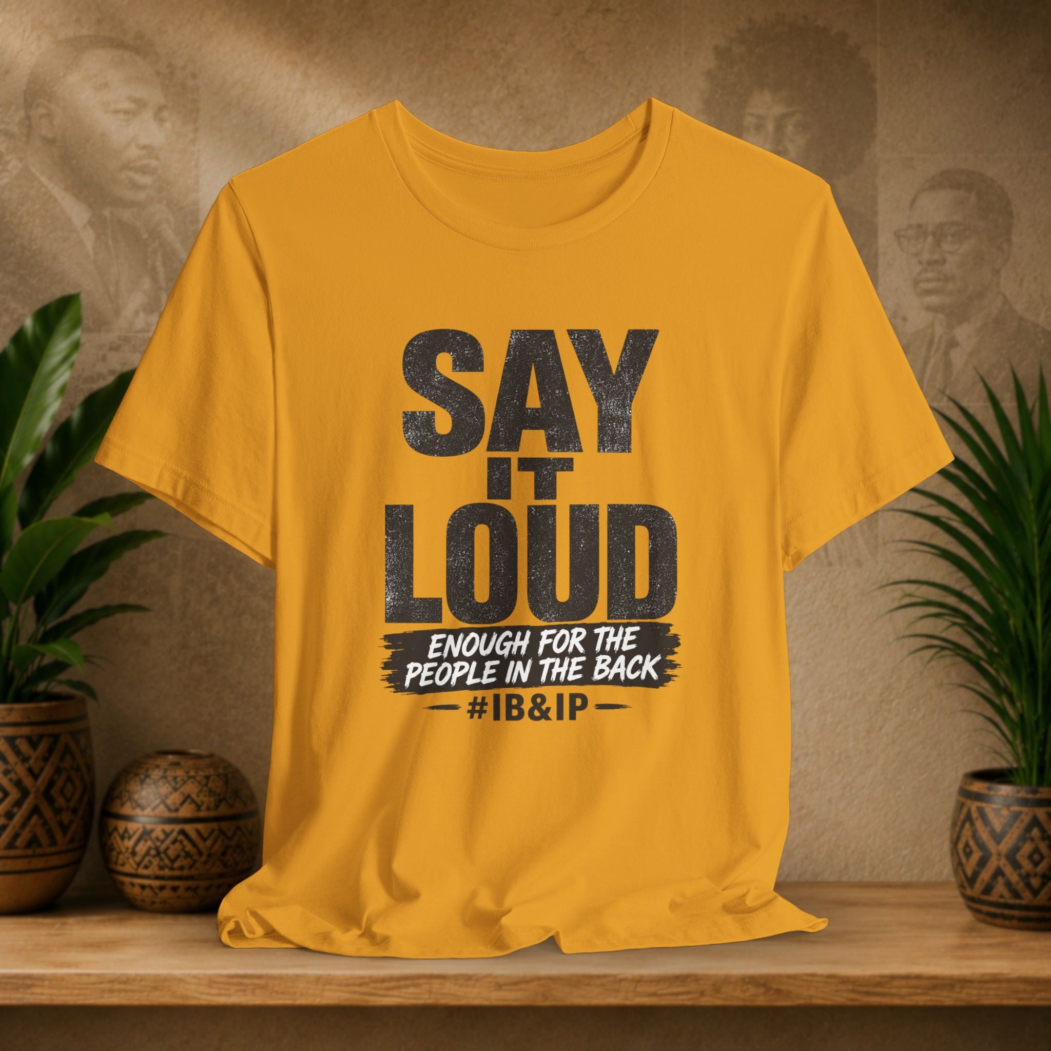Mustard t-shirt with text 'Say it Loud Enough for the People in the Back #IB&IP' on a wooden surface with plants and a textured wall background.