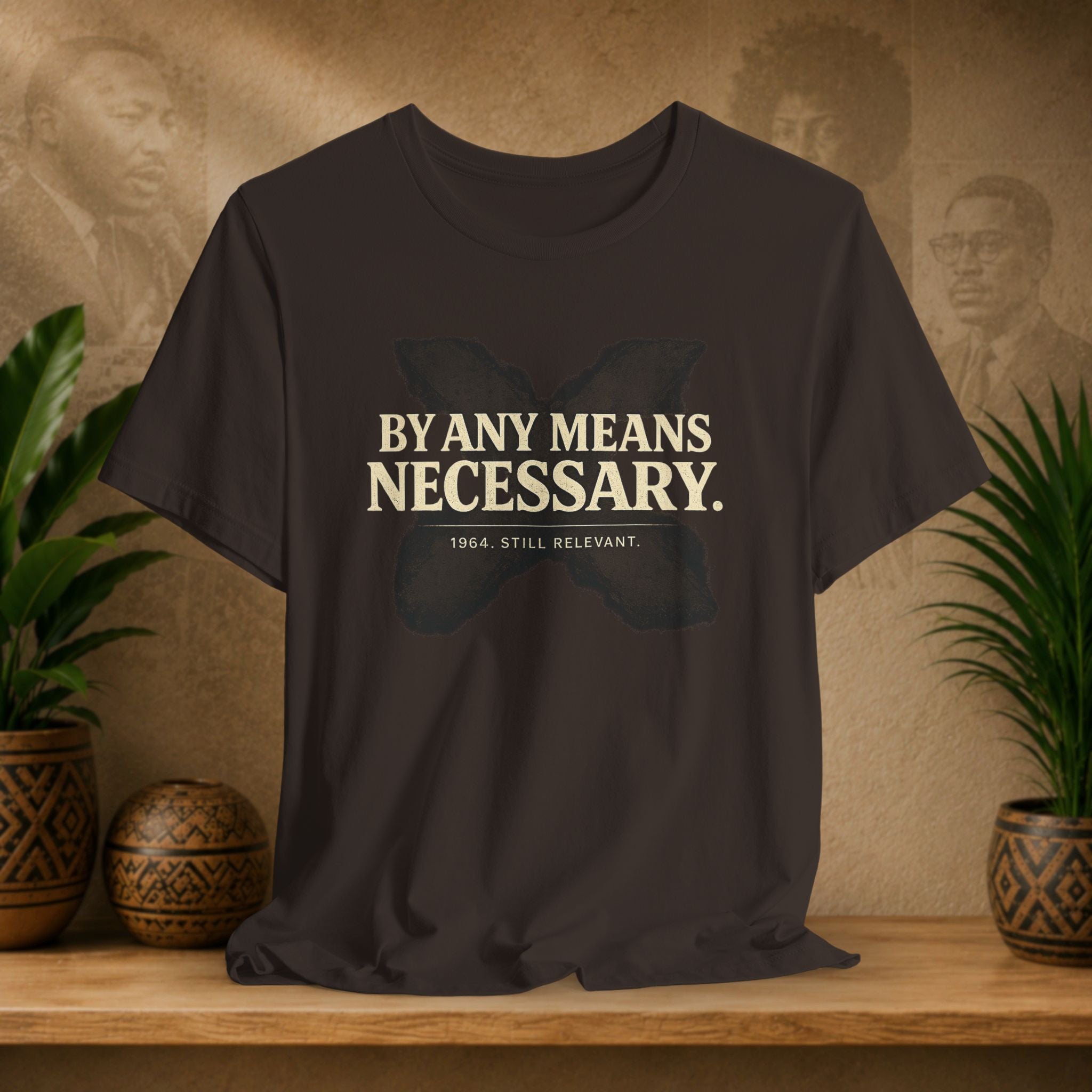 Brown t-shirt with 'By Any Means Necessary' text on a wooden surface with plants and a vintage-style wall in the background.
