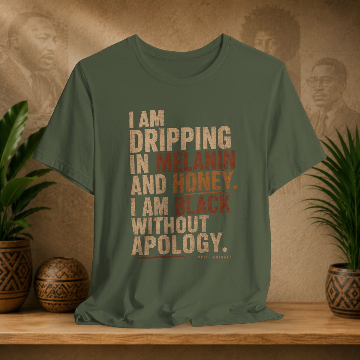 An olive green graphic t-shirt featuring a quote by Nate Goldberg, displayed on a wooden shelf with decorative plants and textured background.