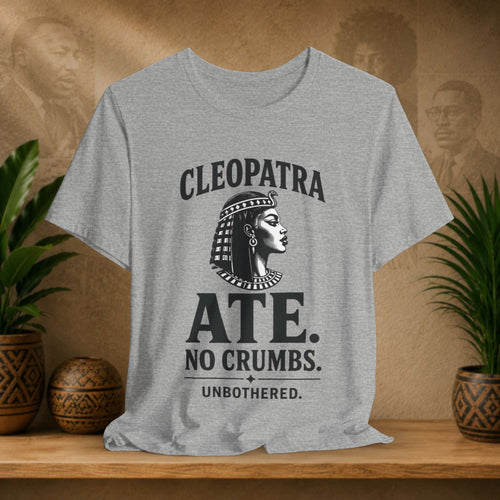 Gray t-shirt with Cleopatra graphic and text on a wooden surface with plants and decorative items.