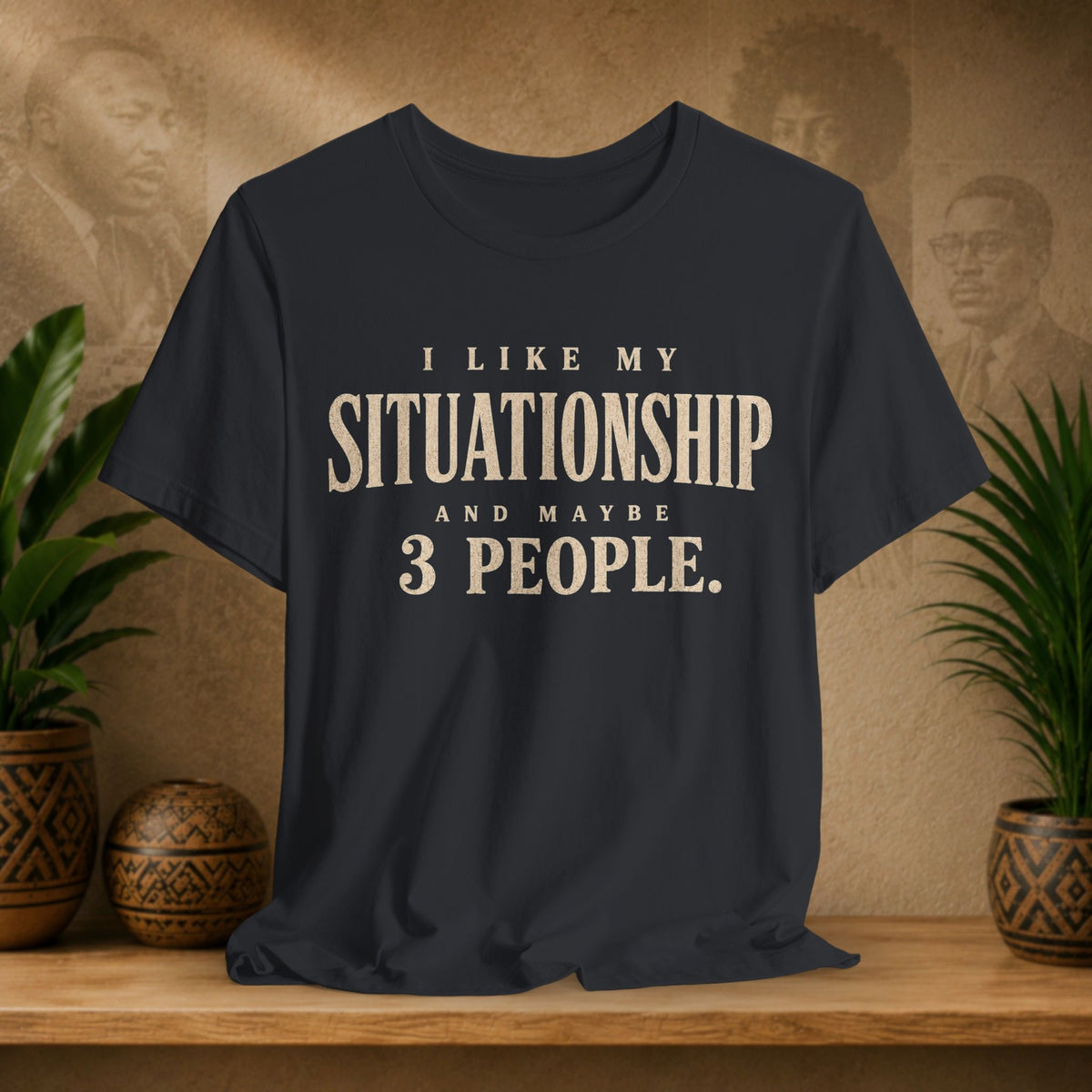 A black graphic t-shirt displayed on a wooden shelf, flanked by potted green plants and decorative ceramic pots against a textured beige background with sepia portraits.