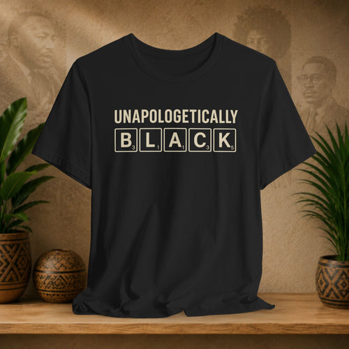 Black t-shirt with 'UNAPOLOGETICALLY BLACK' text on a wooden surface with plants and a textured wall background.