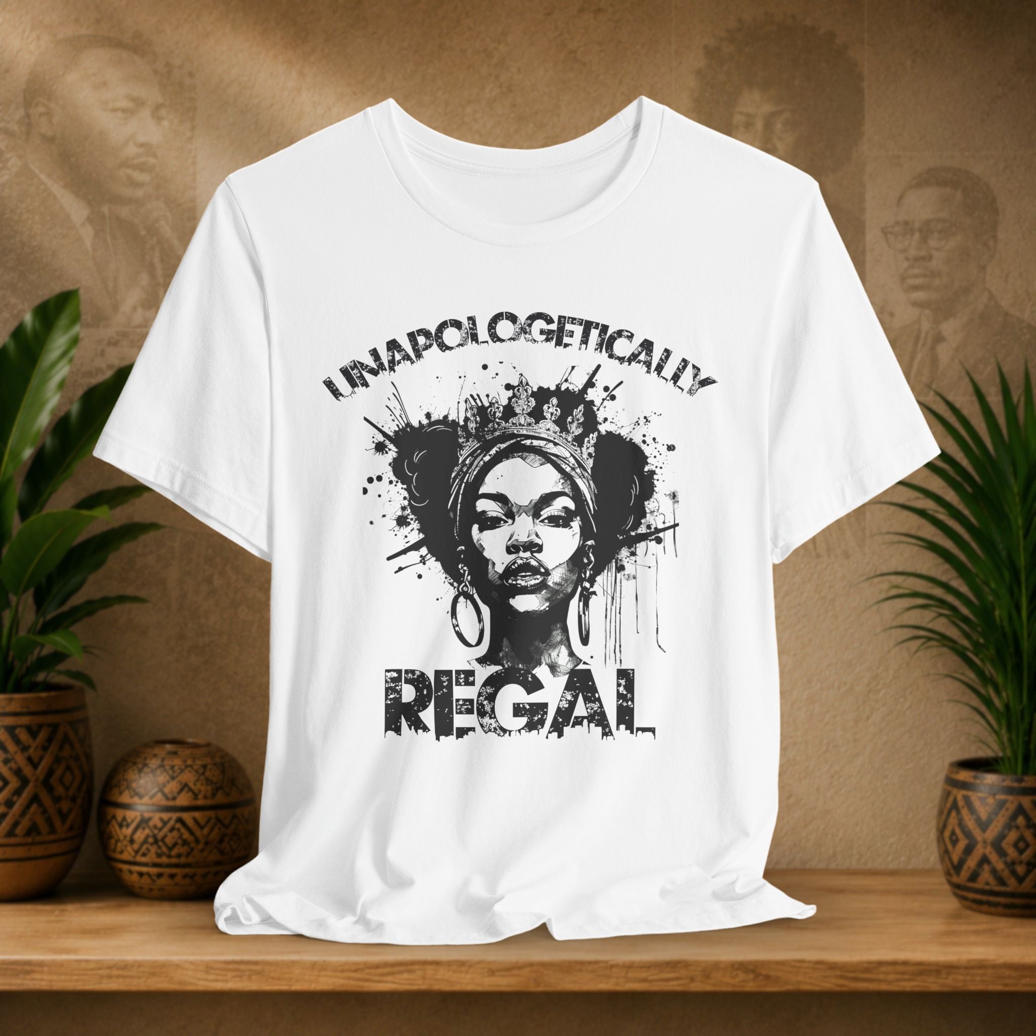 A white short-sleeve graphic T-shirt displayed on a wooden surface, featuring a black distressed print of a woman's portrait with the words 'UNAPOLOGETICALLY REGAL.' The shirt is styled in a warm indoor setting with potted plants and a textured wall in the background.