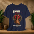 A navy blue graphic t-shirt displayed on a wooden shelf with potted plants in the background. The shirt features a graphic of a hand holding a finger to the lips in front of red lips, with the text 'SHHH.