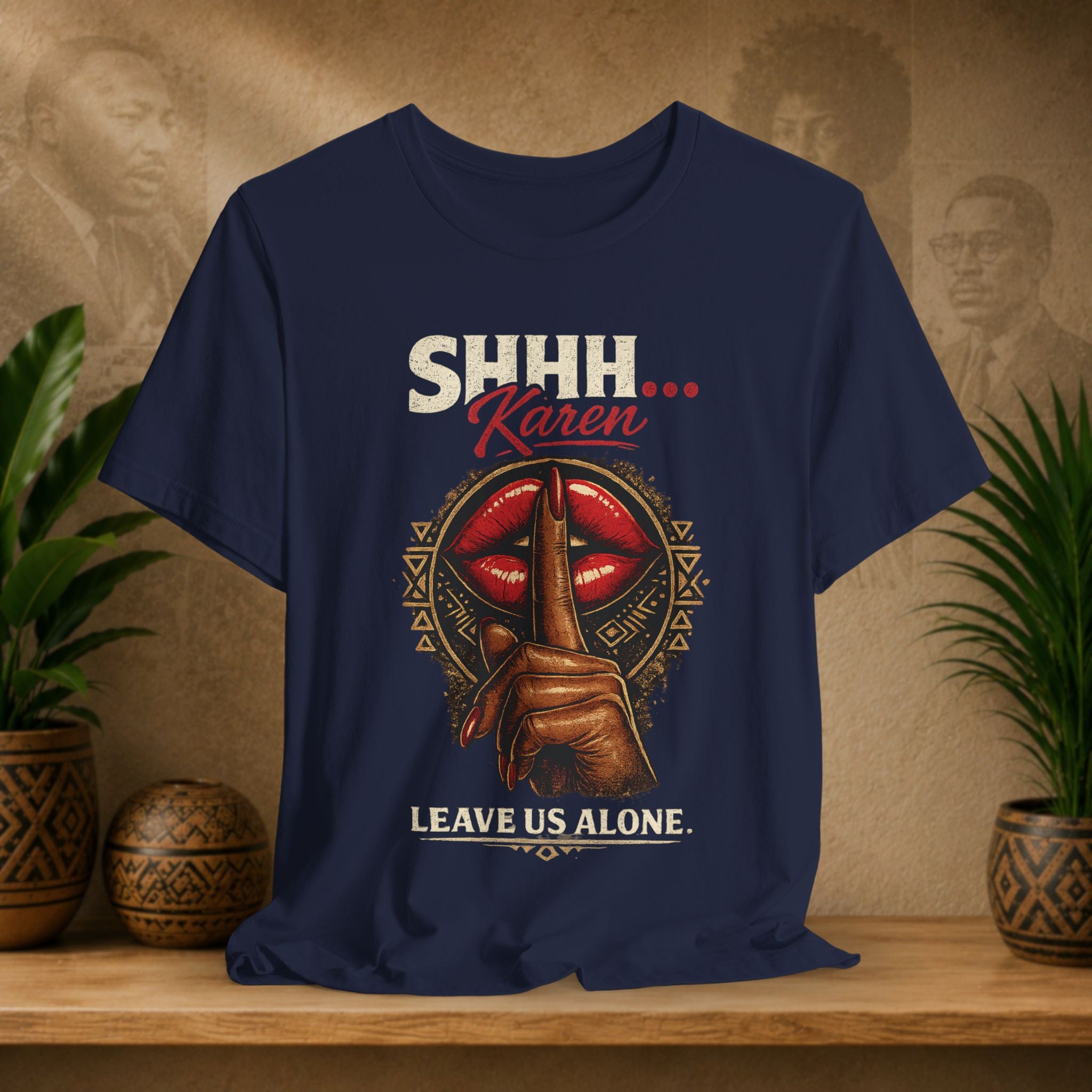 A navy blue graphic t-shirt displayed on a wooden shelf with potted plants in the background. The shirt features a graphic of a hand holding a finger to the lips in front of red lips, with the text 'SHHH.