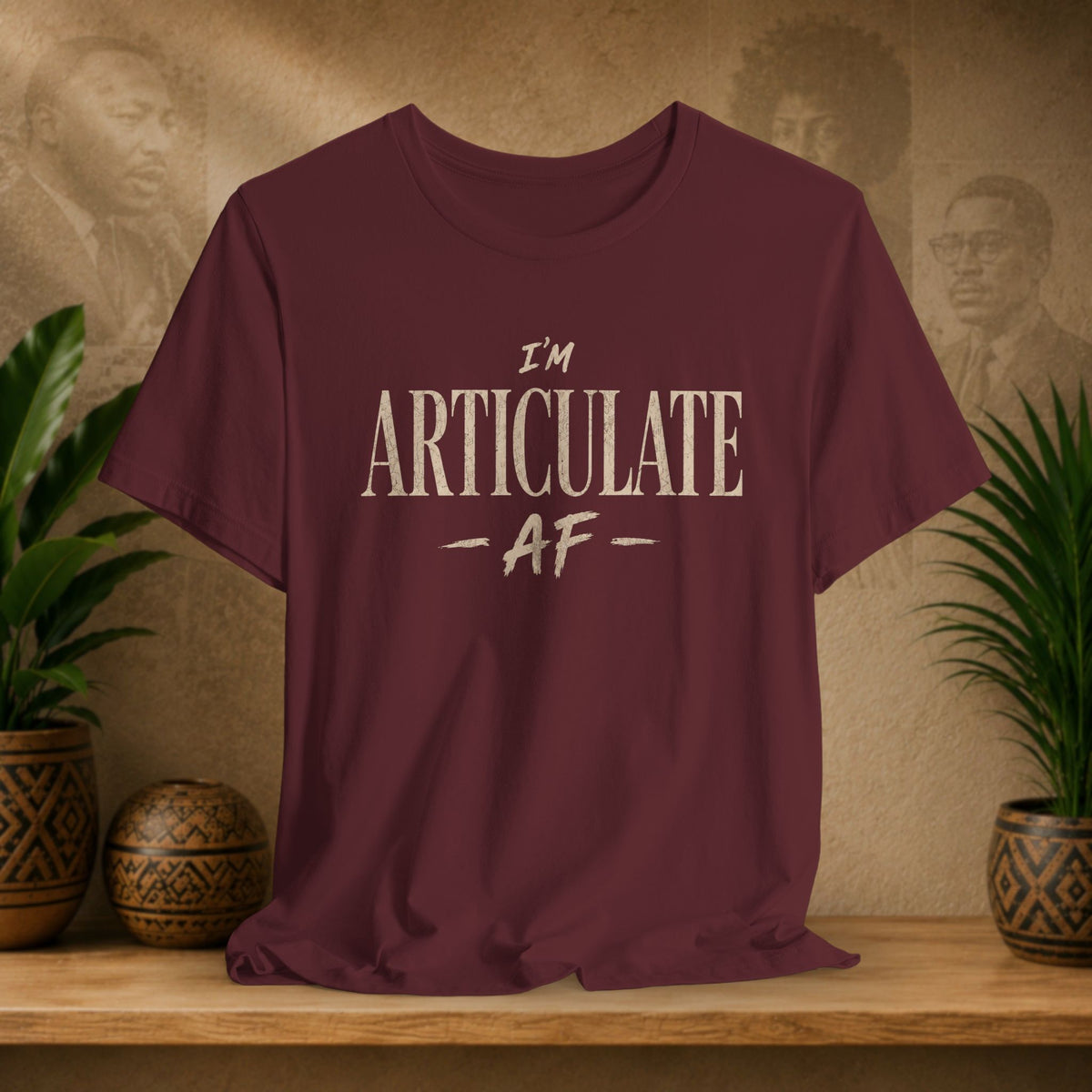 A maroon crewneck t-shirt with the text 'I'M ARTICULATE AF' printed in a distressed beige font, displayed on a wooden shelf with potted plants and a textured background.