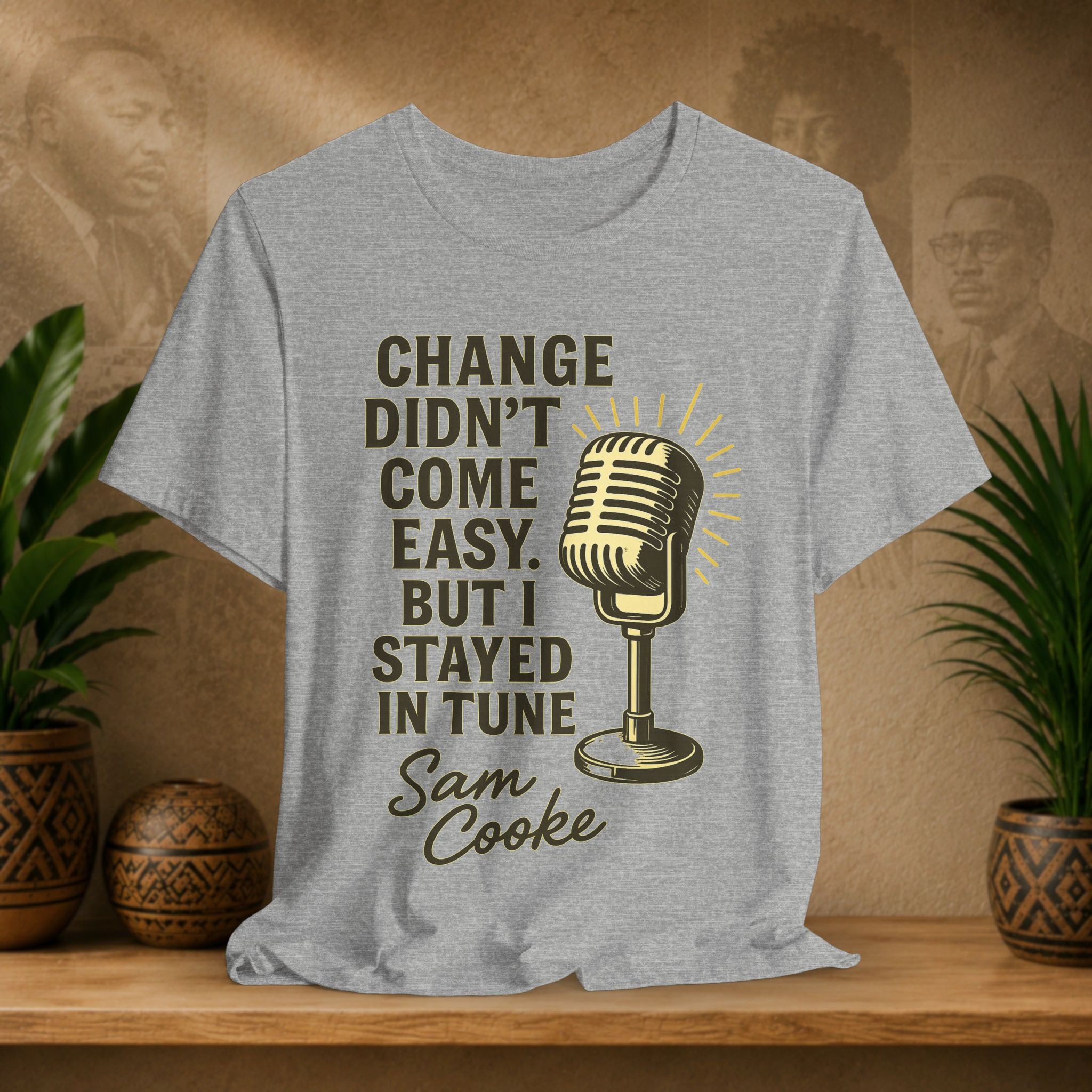 Gray t-shirt with a microphone graphic and text on a wooden surface with plants and a wall in the background
