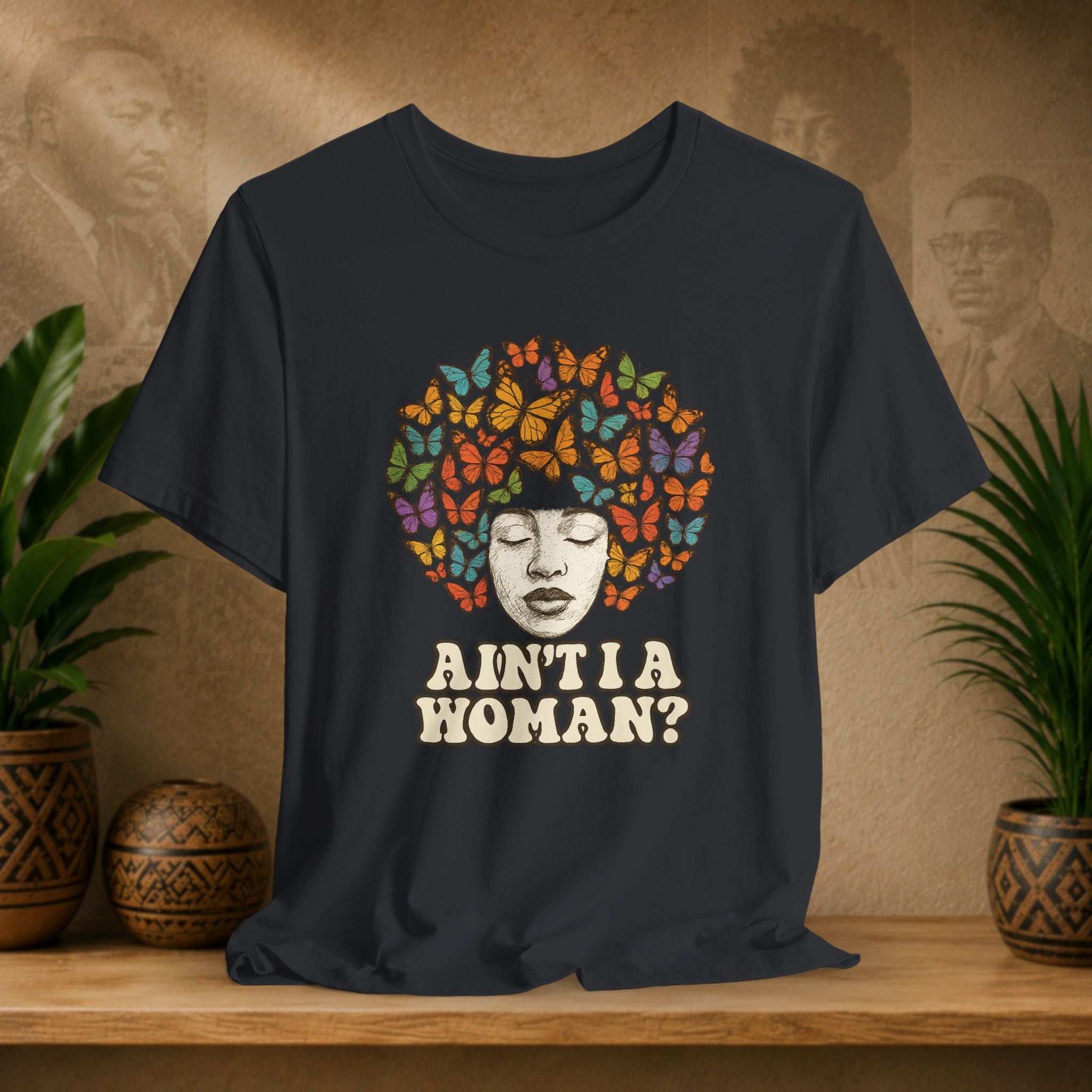 Black t-shirt displayed on a wooden shelf, featuring a graphic of a Black woman's face surrounded by colorful butterflies with the text 'AIN'T I A WOMAN?' in cream. The background shows a warm, textured wall with sepia portraits and potted plants.