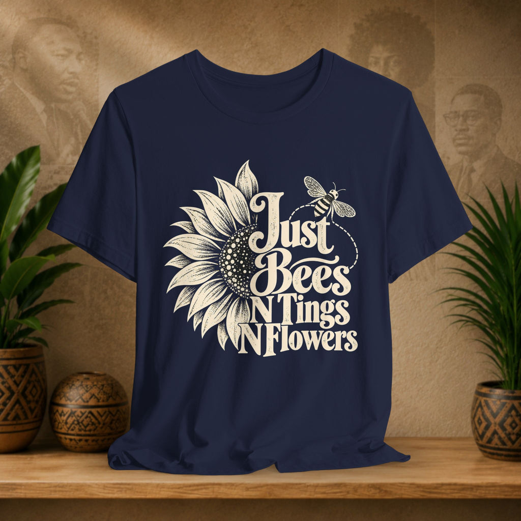 A navy blue short-sleeve t-shirt with a white graphic print of a sunflower, a bee, and the text 'Just Bees N Tings N Flowers', displayed on a wooden shelf with potted plants and a textured background.
