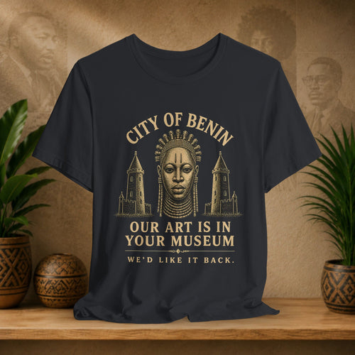 Black t-shirt with gold text and graphic of a face and towers on a wooden surface with plants and decorative items.