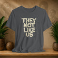 Gray t-shirt with 'THEY NOT LIKE US' text on a wooden surface with plants and a textured wall background