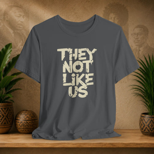 Gray t-shirt with 'THEY NOT LIKE US' text on a wooden surface with plants and a textured wall background