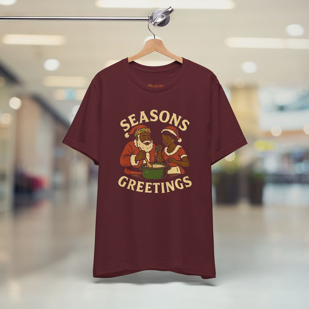 Maroon t-shirt with 'Seasons Greetings' graphic on a hanger in a store setting