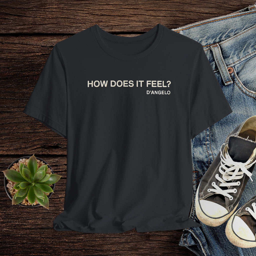 Black t-shirt with 'HOW DOES IT FEEL? D'ANGELO' text on a wooden surface with jeans and sneakers.